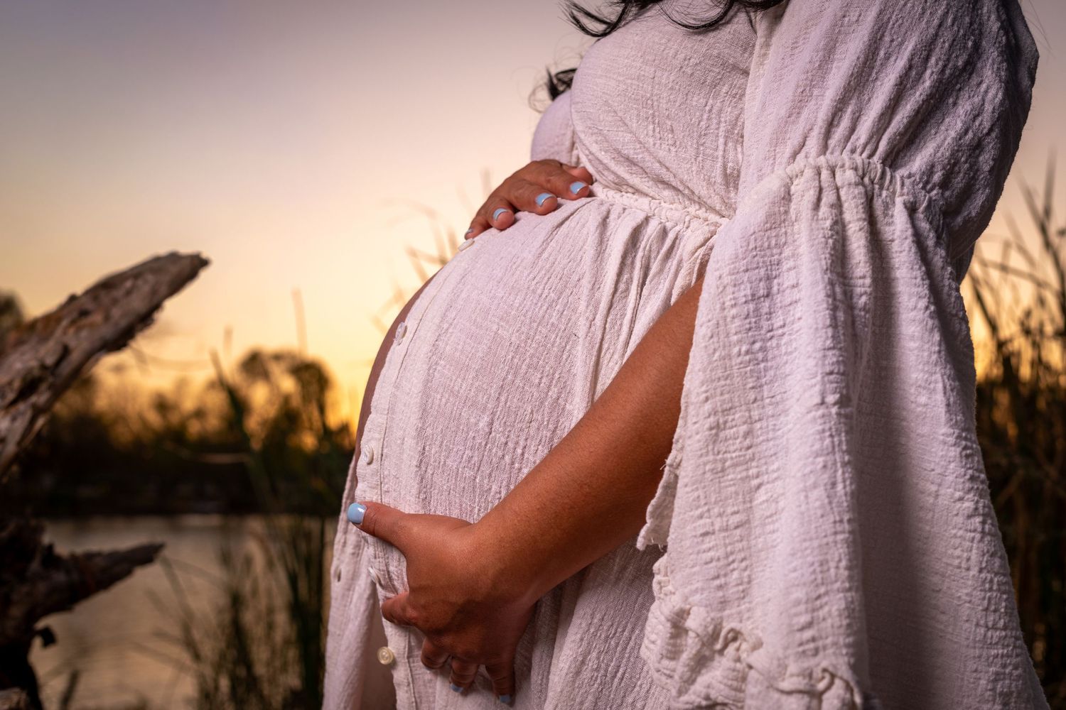 A pregnant figure in a flowing white dress cradles their belly during sunset by a lake.