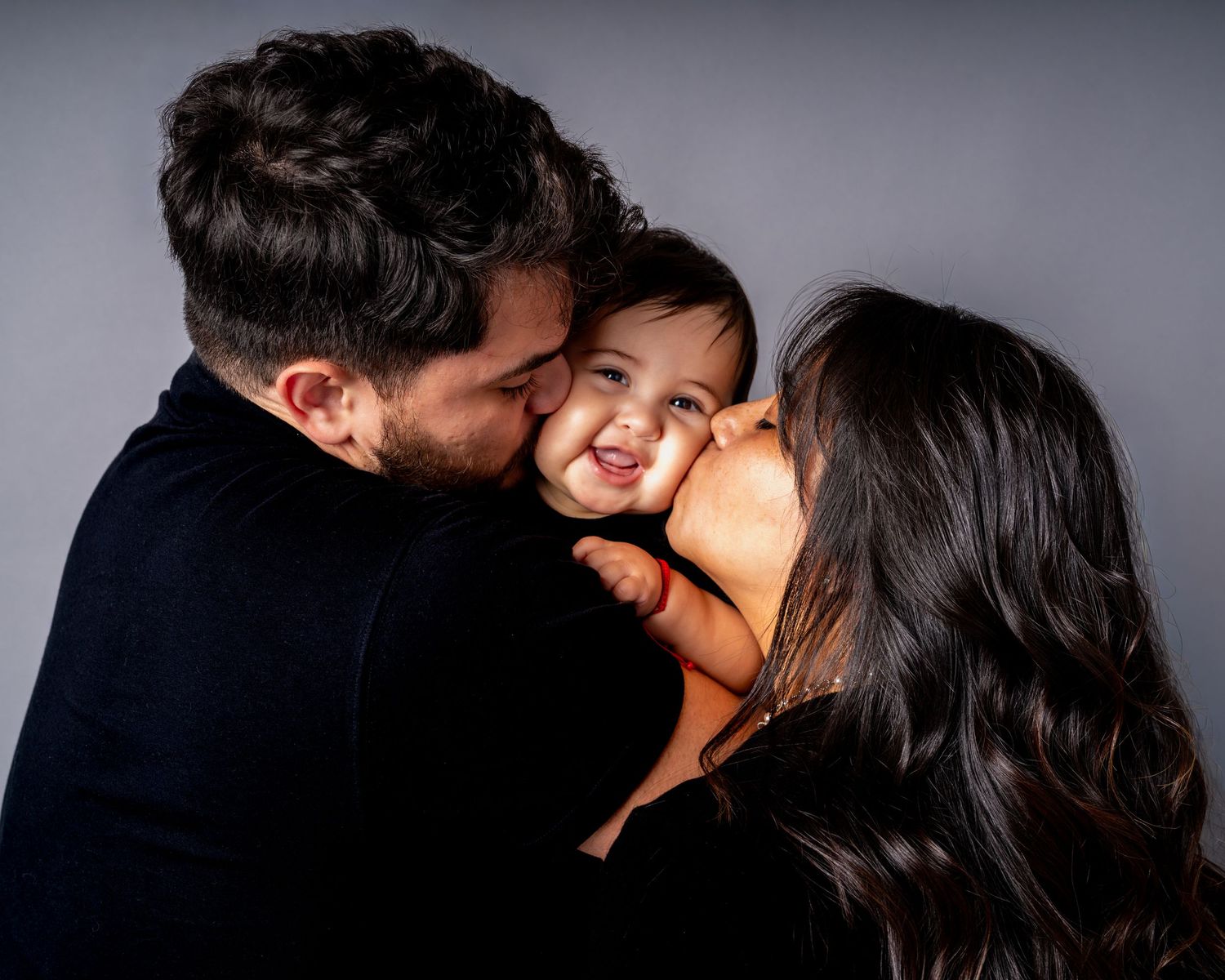A joyful family moment captured in studio lighting as parents kiss their smiling child's cheeks.