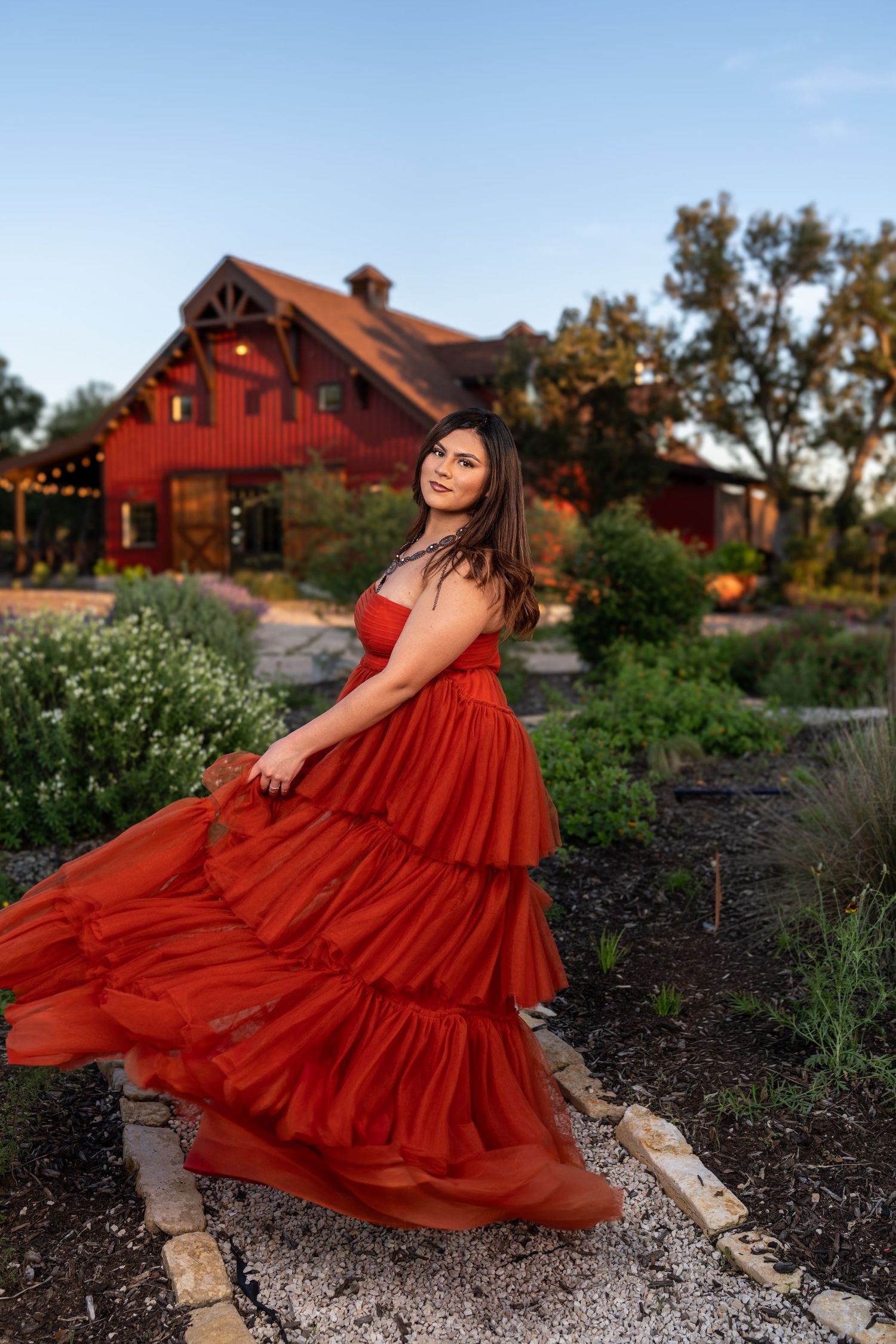 Woman in flowing red tiered gown twirling in garden at dusk with rustic red barn in background.
