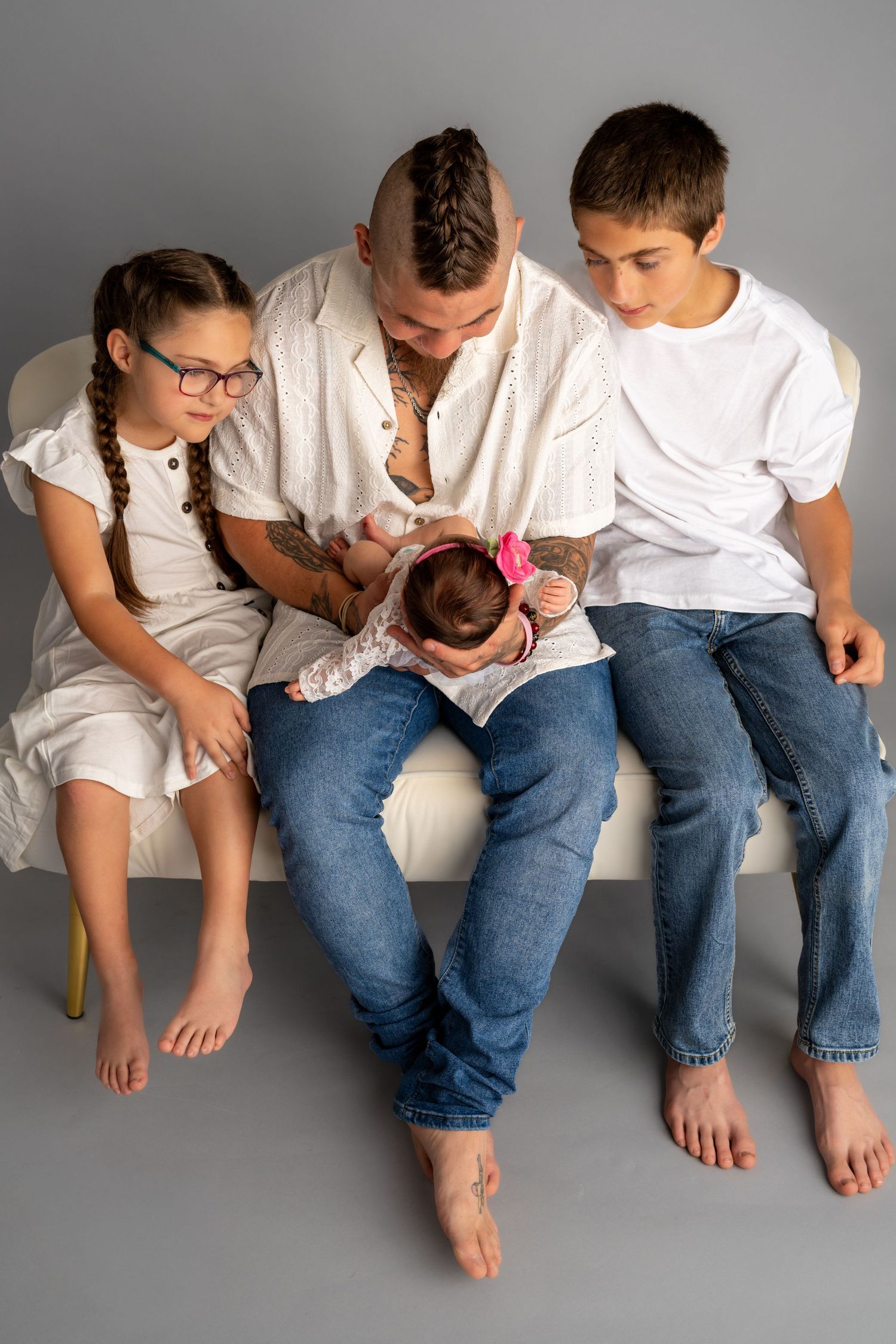 Father with mohawk tattoos sits on bench with three children, including newborn baby, in white outfits on gray background.