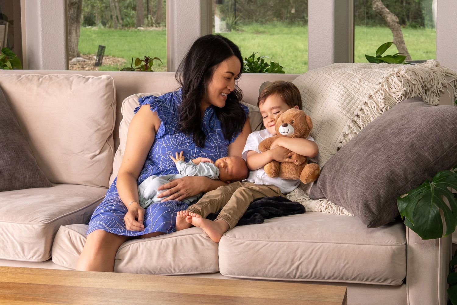Mother in blue dress sitting on couch with newborn baby and toddler holding teddy bear.
