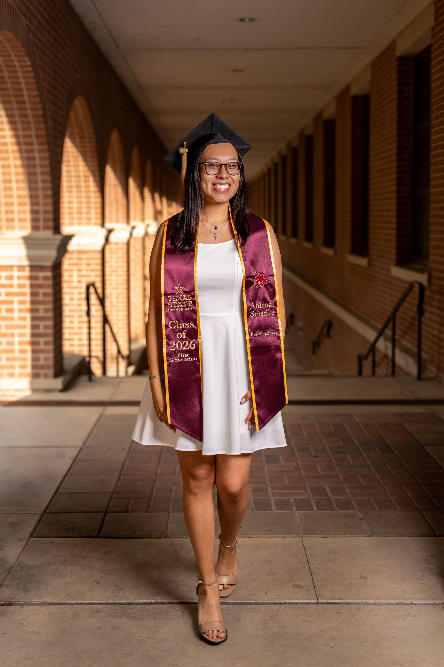 A smiling graduate poses in white dress, maroon stole reading Class of 2026, and cap in a brick building hallway.