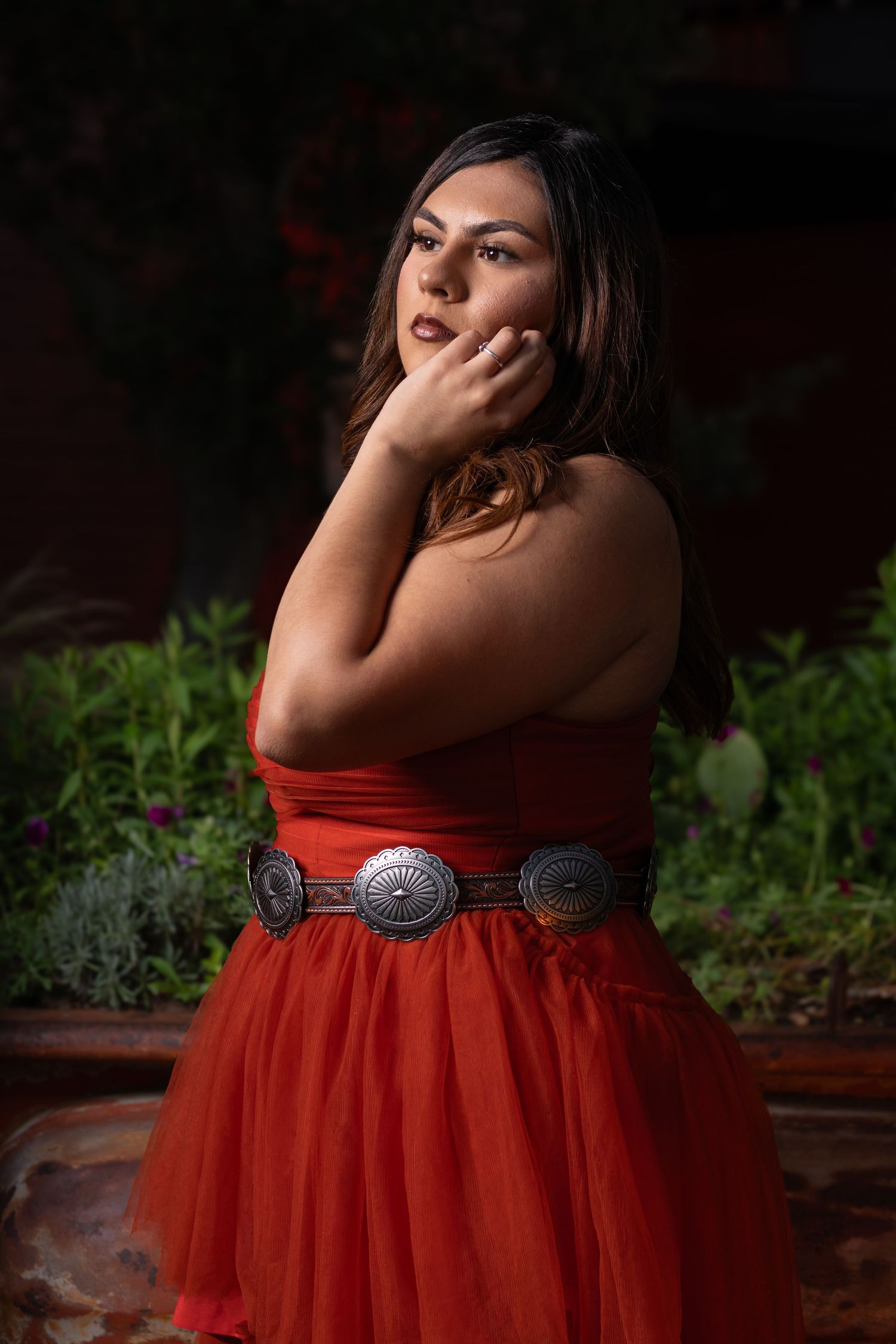 Woman in red strapless dress with silver concho belt posing outdoors in garden setting.
