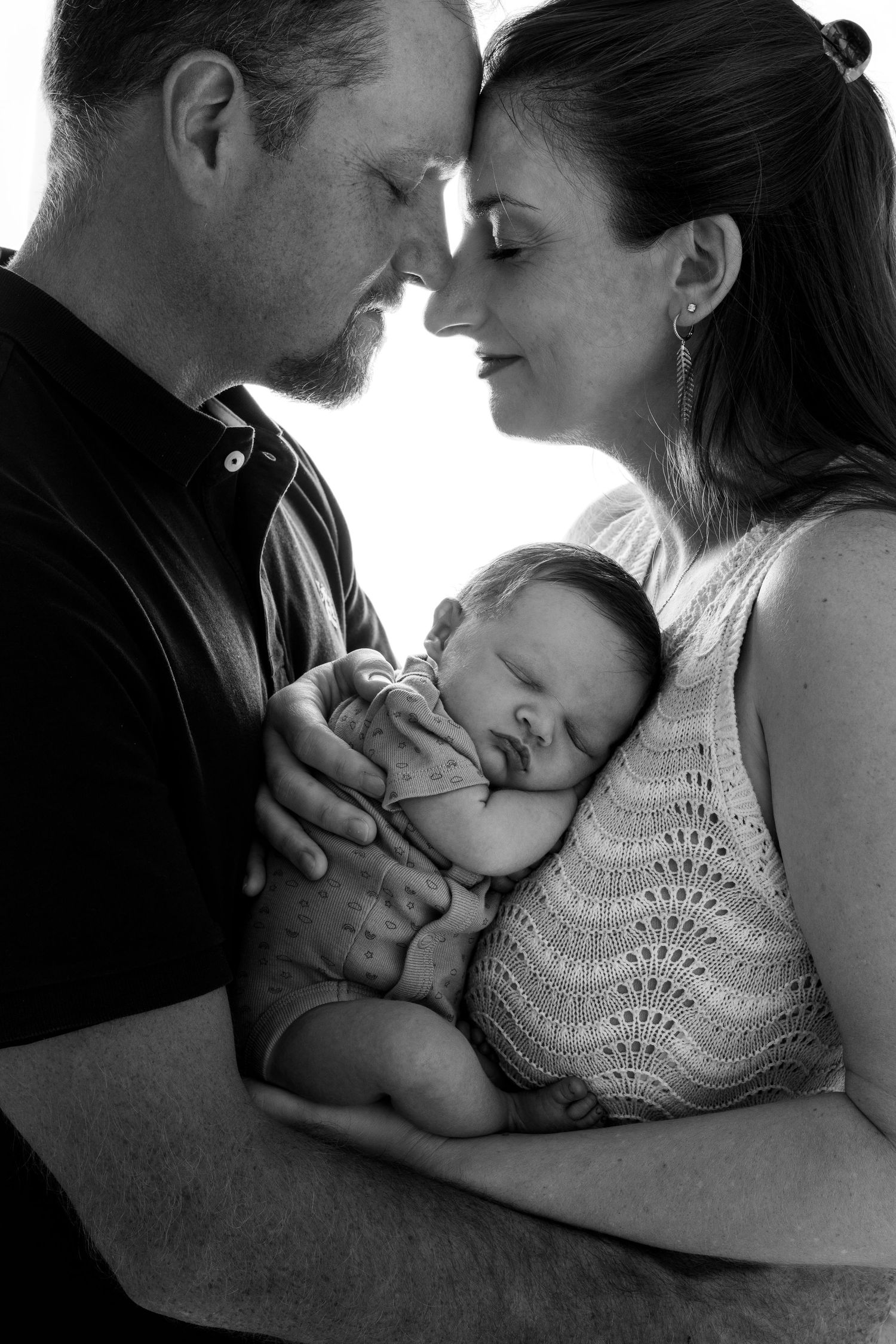 Black and white photo of parents tenderly holding their sleeping newborn baby, foreheads touching in an intimate family portrait.