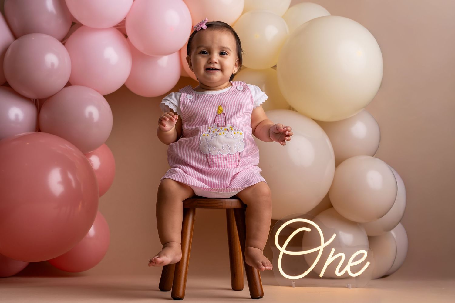 Baby girl in pink romper sits on stool surrounded by pink and cream balloons celebrating her first birthday.
