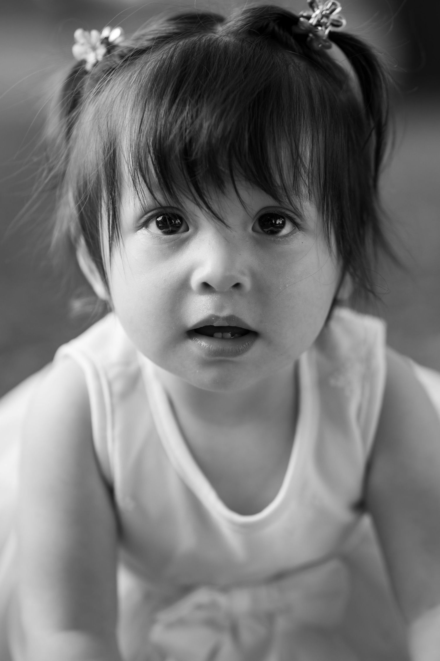 Black and white portrait of a baby girl with pigtails wearing a white dress, looking up with wide curious eyes.