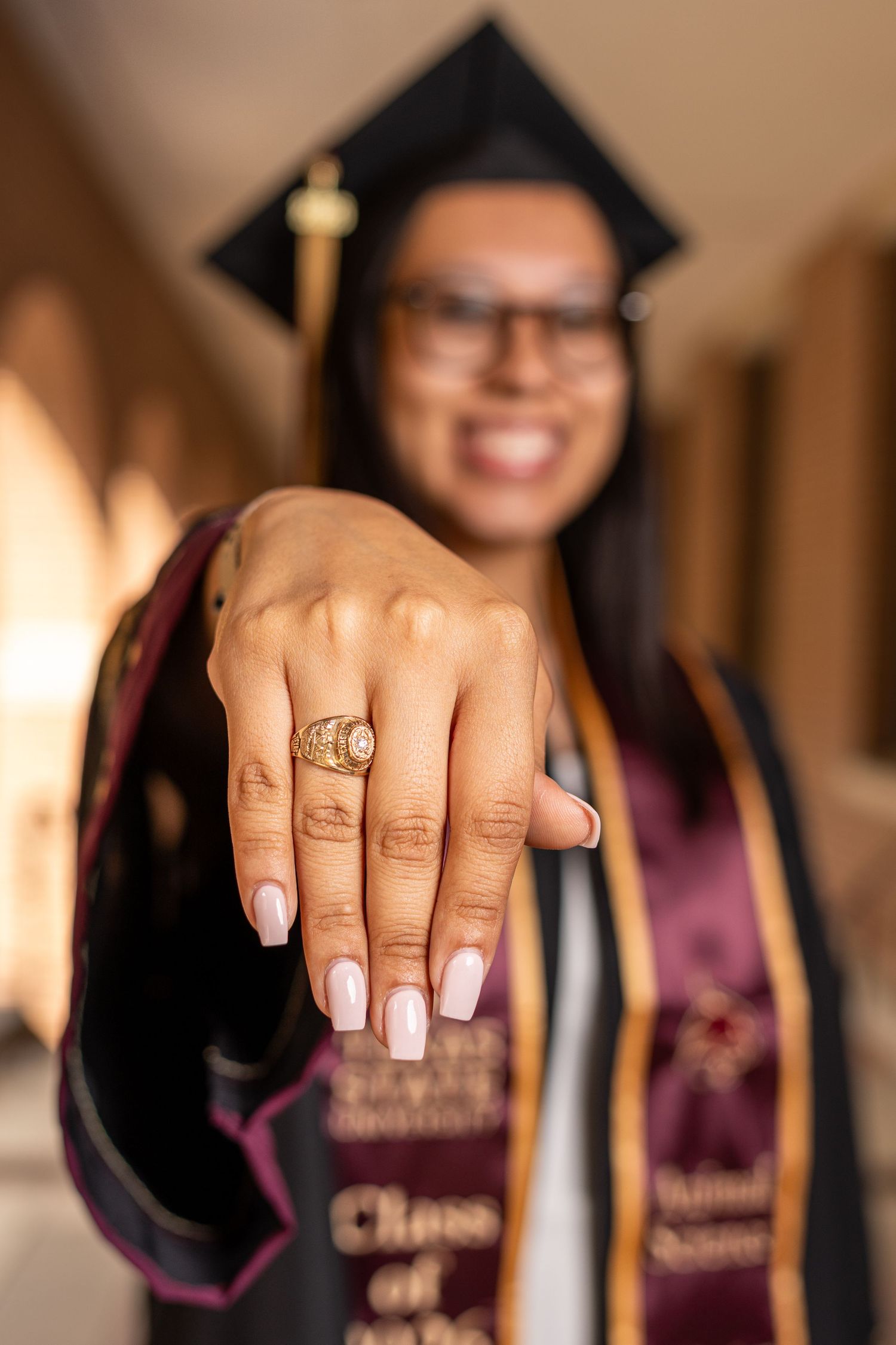 Smiling graduate in cap and gown proudly displays gold class ring with pink manicured nails at graduation ceremony.