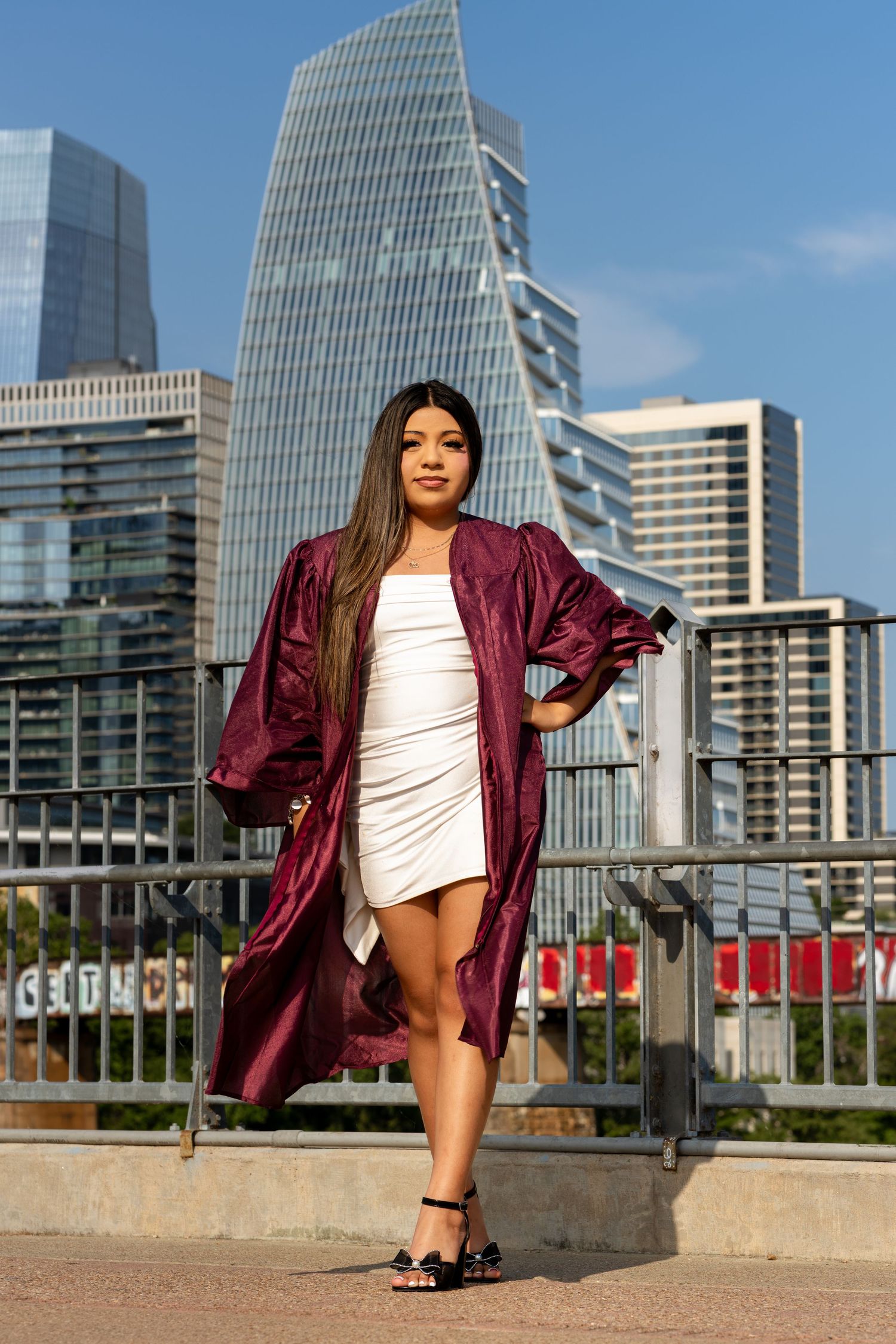 A young woman in a maroon graduation gown and white dress poses confidently in front of a modern city skyline.