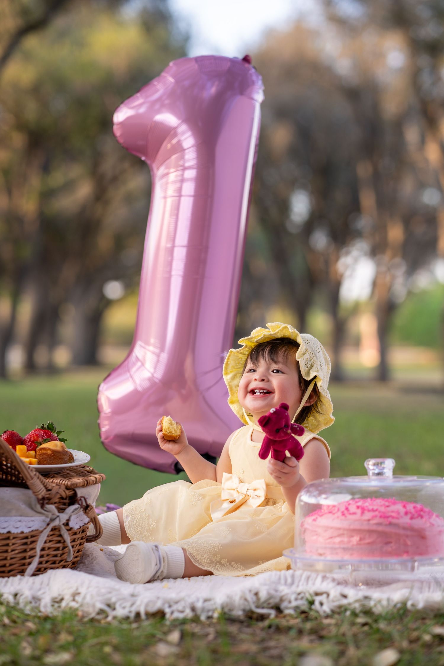 Smiling baby girl in yellow dress celebrates first birthday with pink number one balloon and cake outdoors.