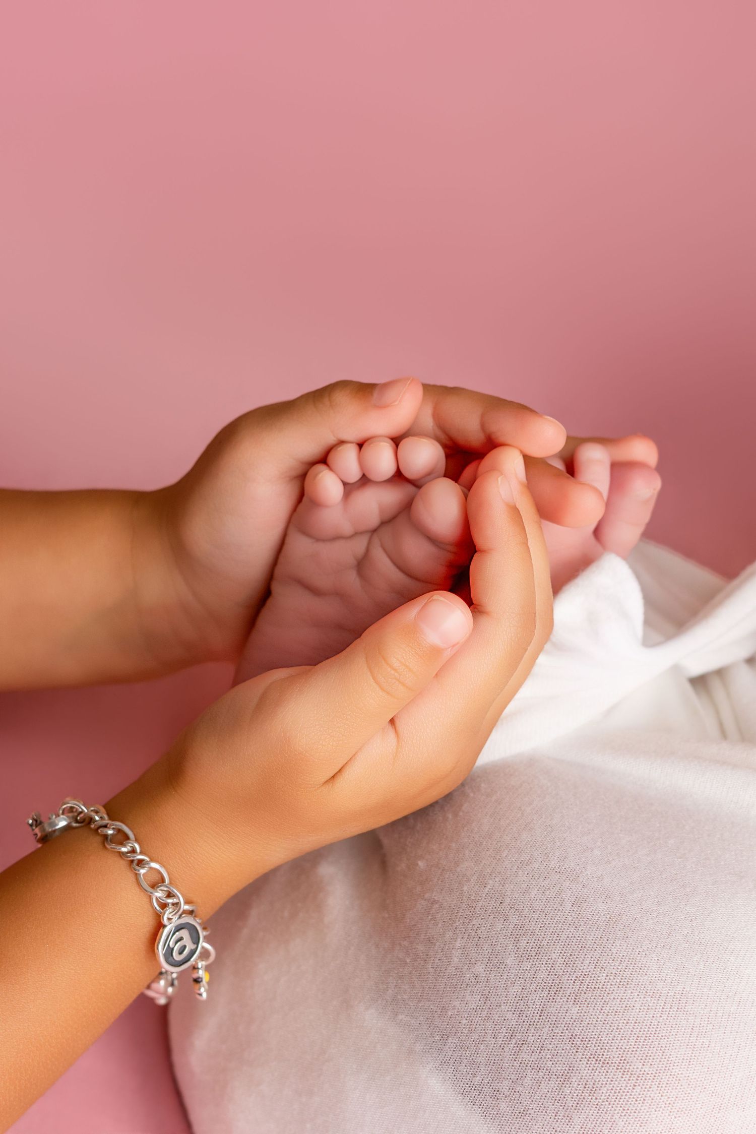Child's hands with a silver charm bracelet gently cradle a newborn baby's tiny feet against a soft pink background.