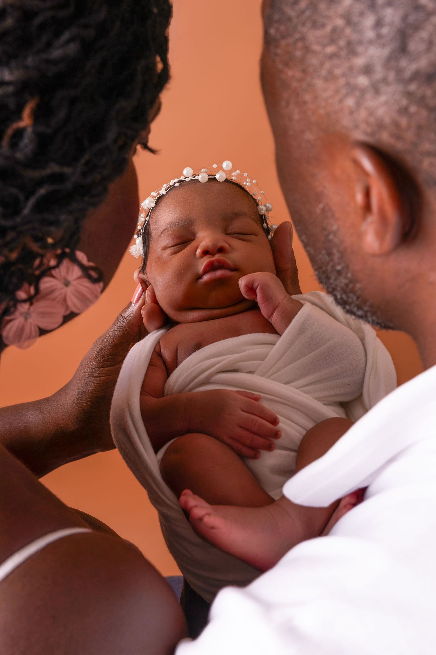 Two parents gently cradle a sleeping newborn wrapped in a cream swaddle and wearing a pearl headband in a warm newborn photo.