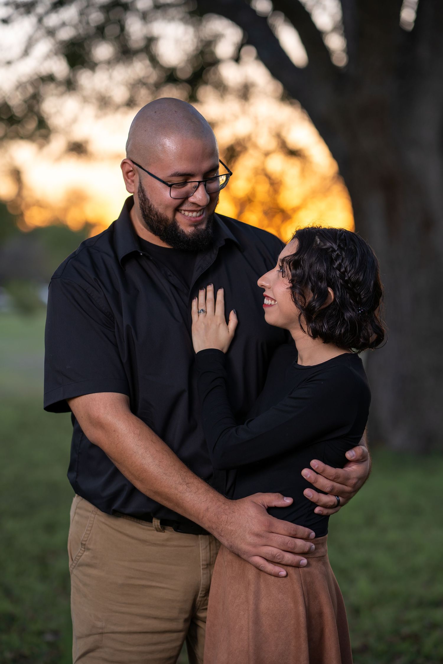 A couple shares an intimate moment embracing and laughing together at sunset in a park setting.