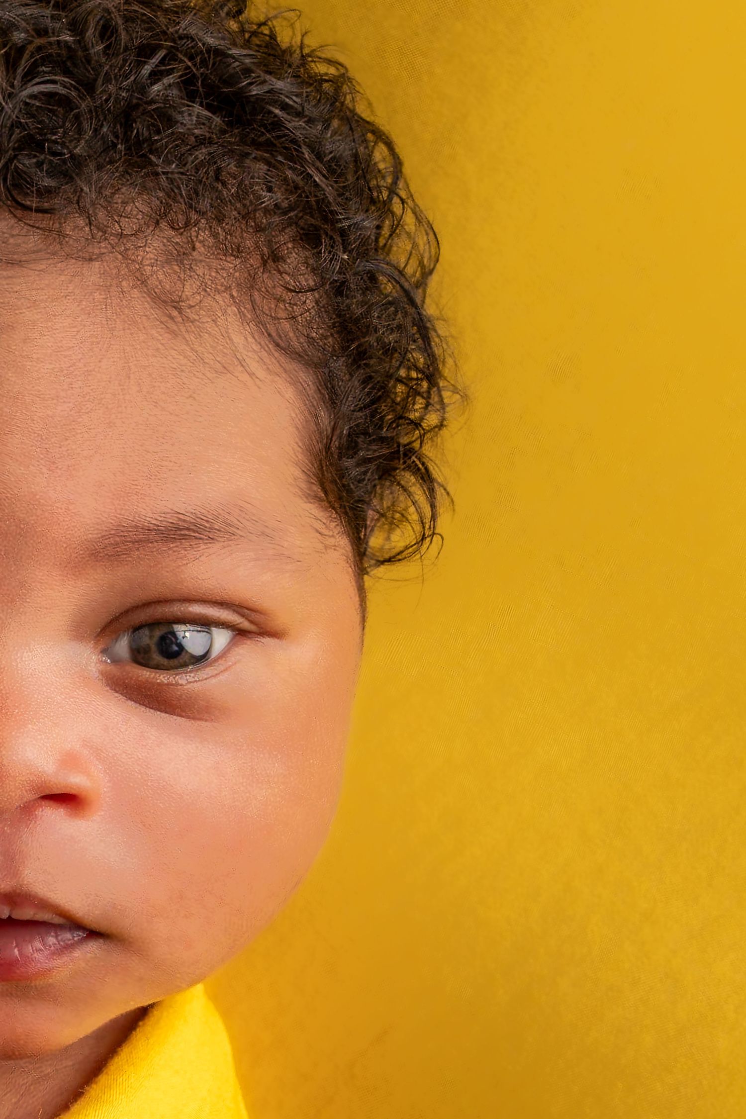 Close-up portrait of a young child with curly hair and brown eyes against a bright yellow background.