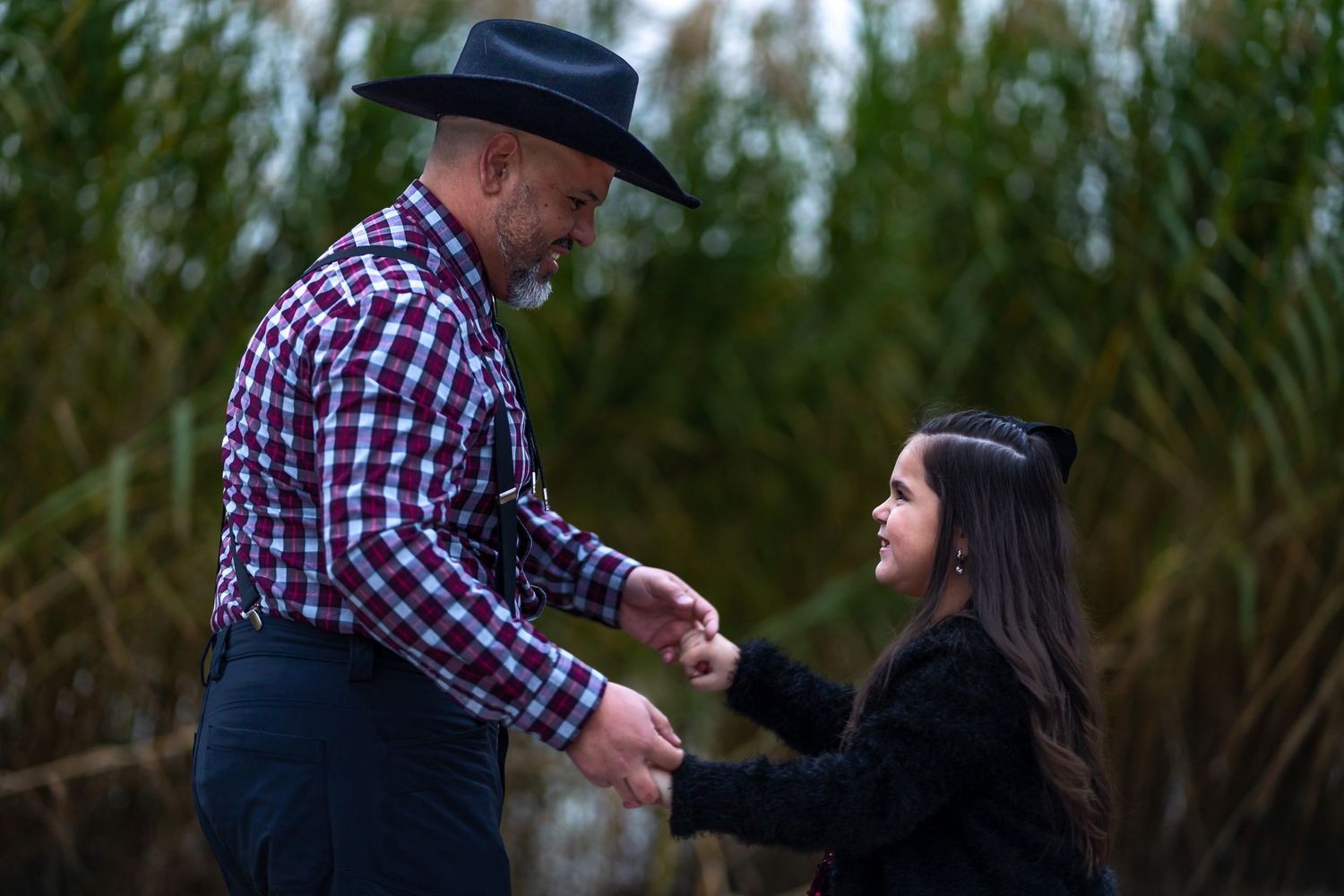 A person in a plaid shirt and hat interacts with someone in a dark outfit against a natural outdoor background.