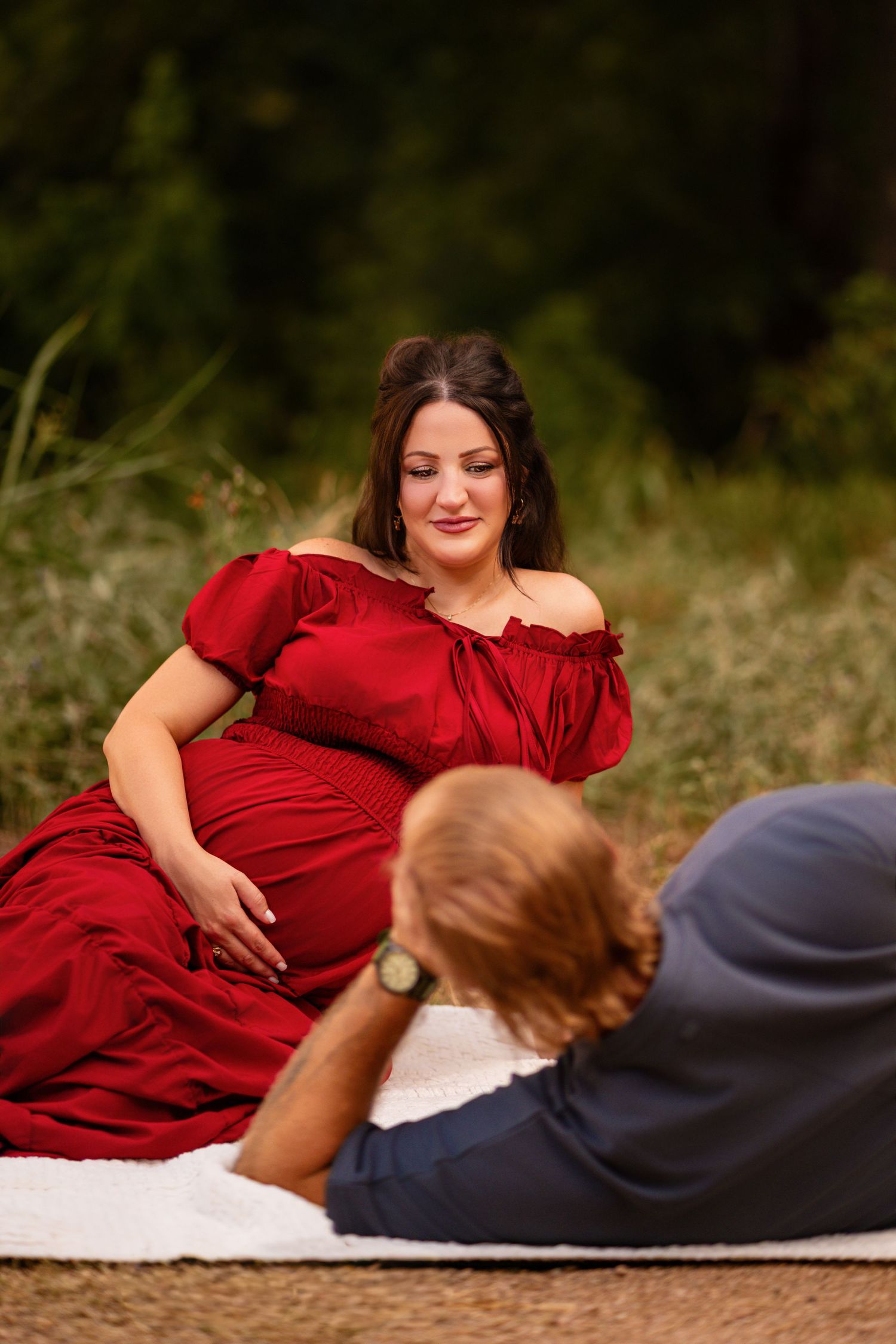Pregnant woman in red off-shoulder gown sits outdoors while partner reclines nearby during a maternity photoshoot.