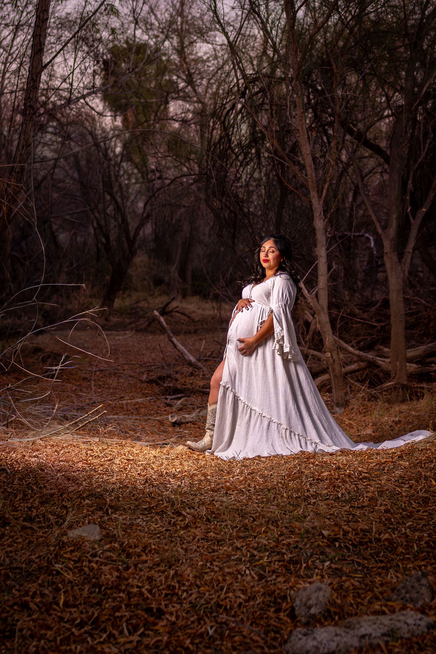 A figure in a flowing white dress poses gracefully among autumn leaves and bare trees in a moody forest setting.