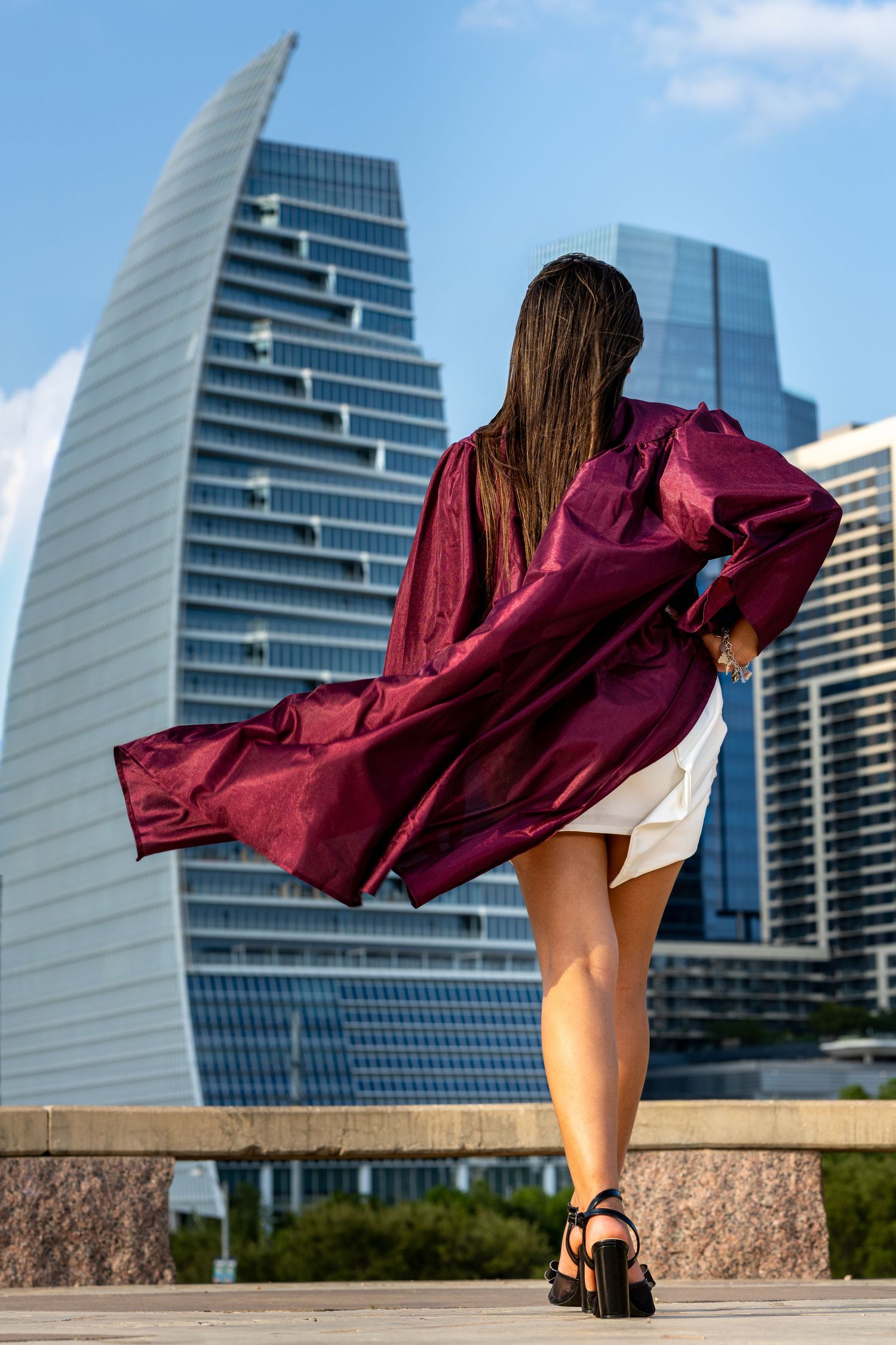 Young woman in burgundy graduation gown and white skirt poses before modern glass skyscrapers on a sunny day.