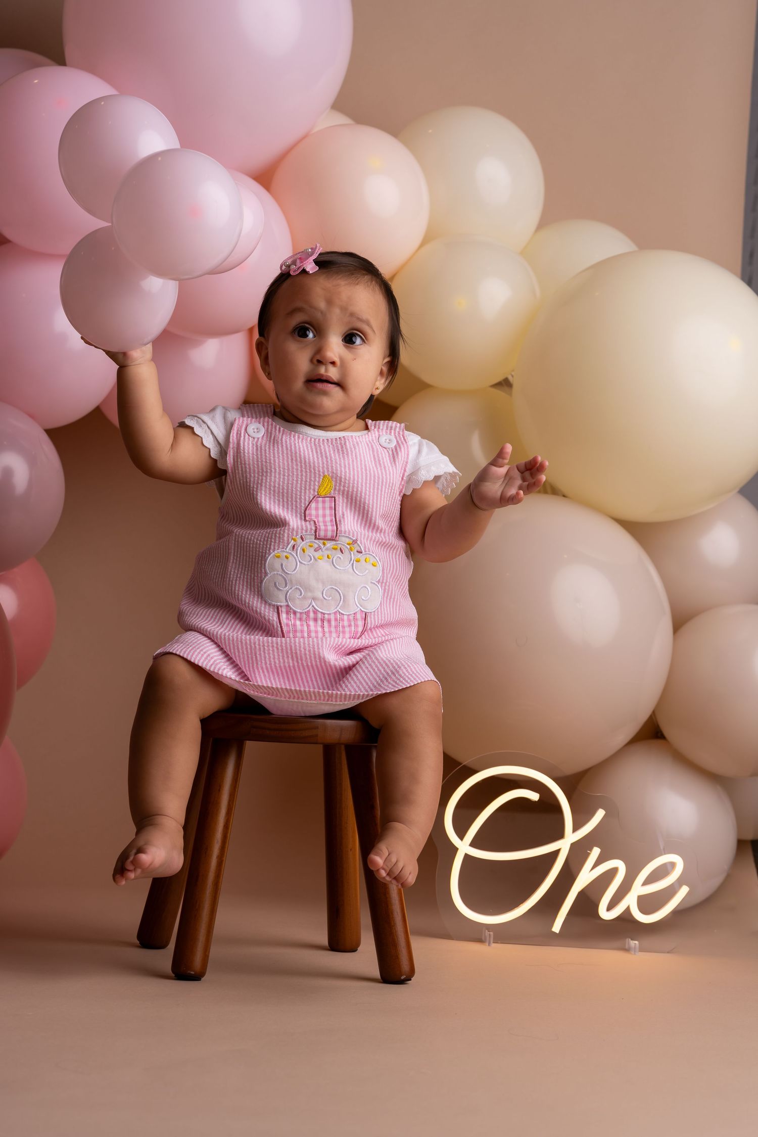 Baby in pink outfit sits on wooden stool against pink and cream balloon backdrop for first birthday photoshoot.
