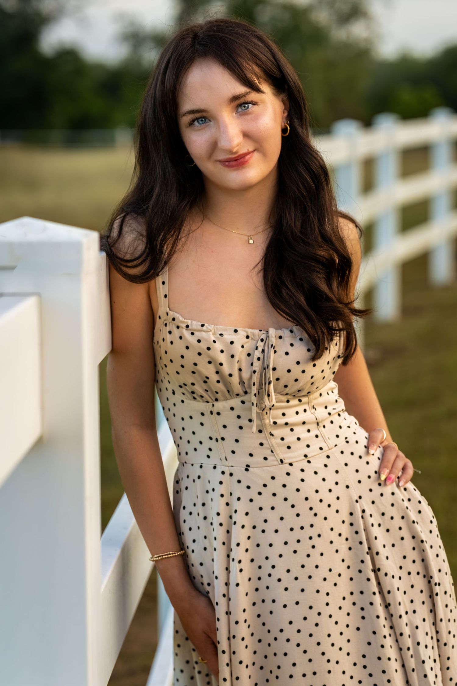 Young woman in polka dot sundress leaning against white fence outdoors.