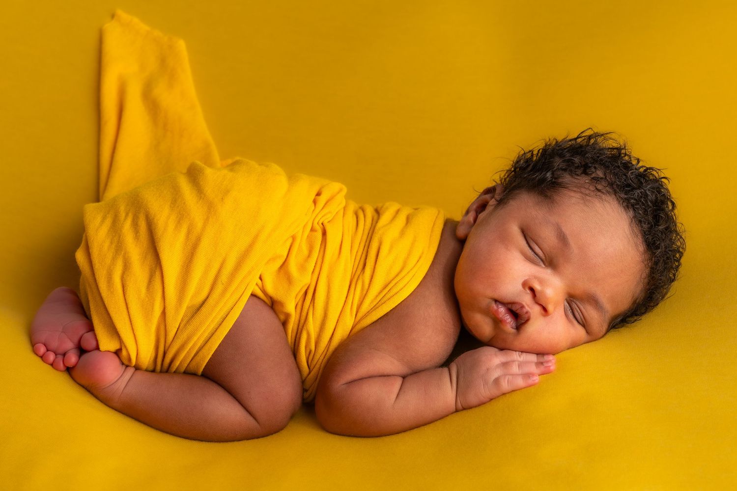 Sleeping newborn baby wrapped in yellow fabric on a yellow background in a peaceful pose.