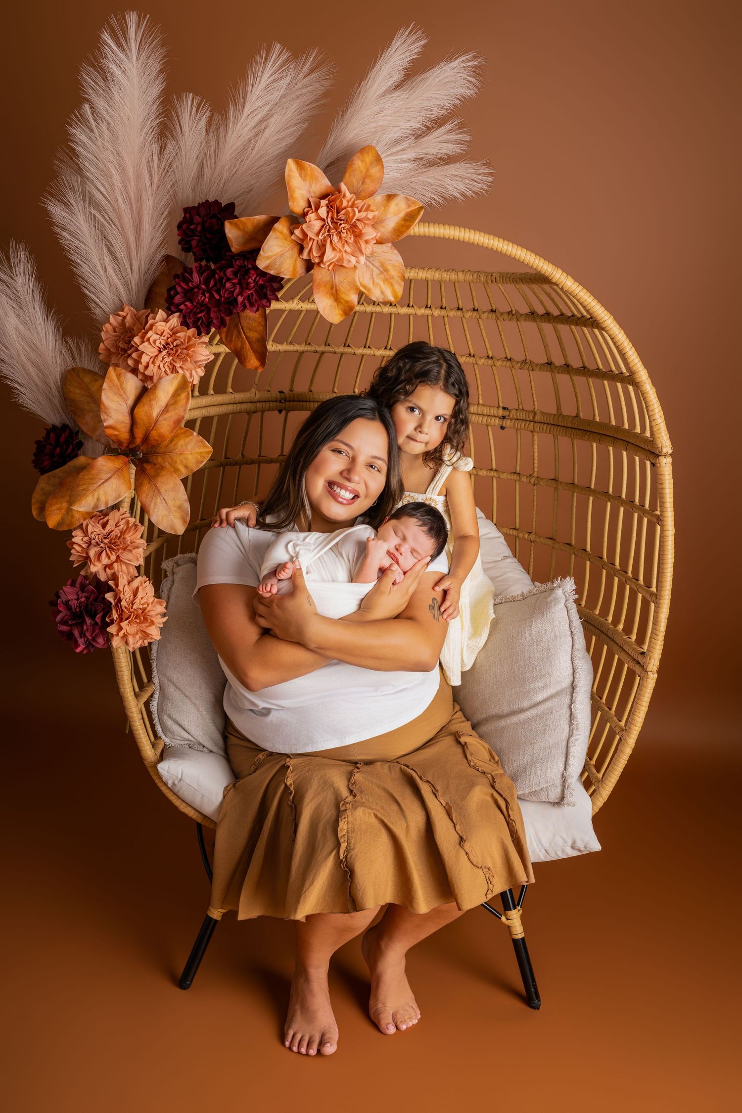 Smiling mother in a rust skirt holds a newborn while a young girl leans on her in a rattan egg chair with boho pampas and dried flowers.