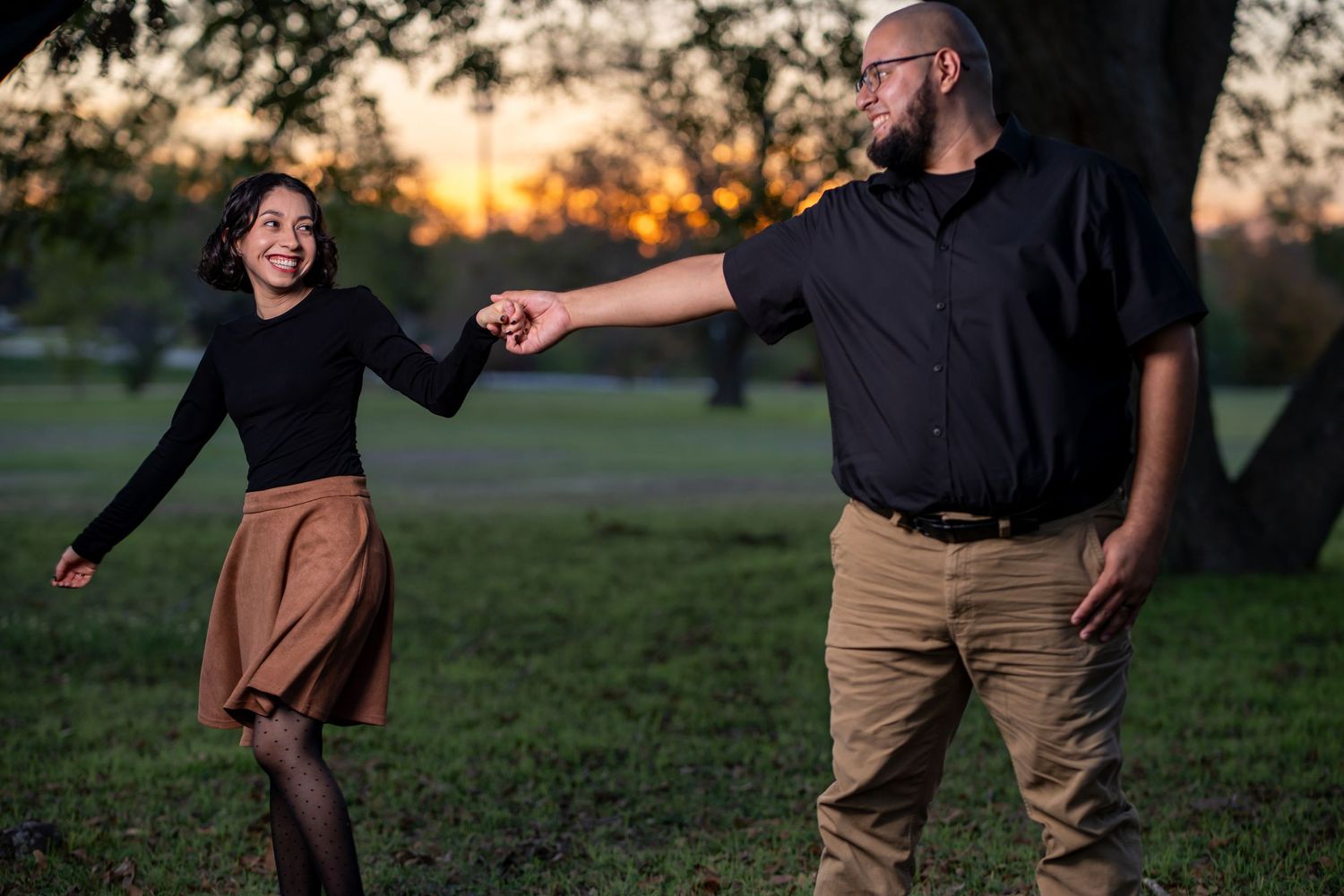 A joyful couple dances together in a park at golden hour with trees in the background.