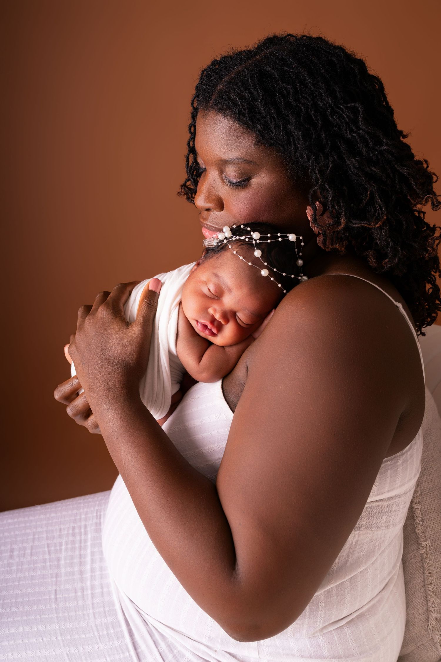 A mother with braided hair cradles her sleeping newborn, who wears a pearl headband, in a tender portrait against a warm brown backdrop.