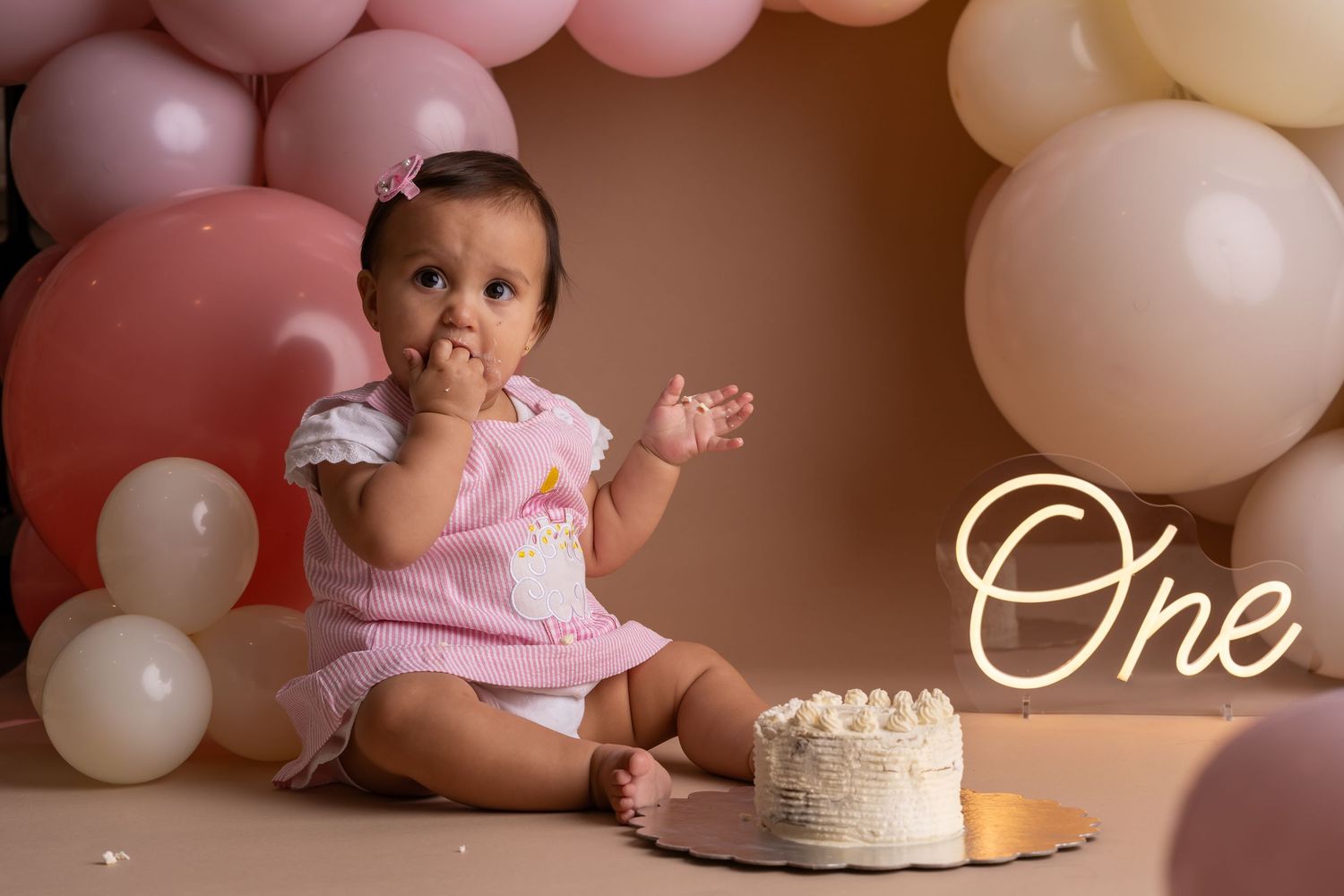 Baby girl in pink dress smashes first birthday cake surrounded by pink and white balloons with glowing One neon sign.