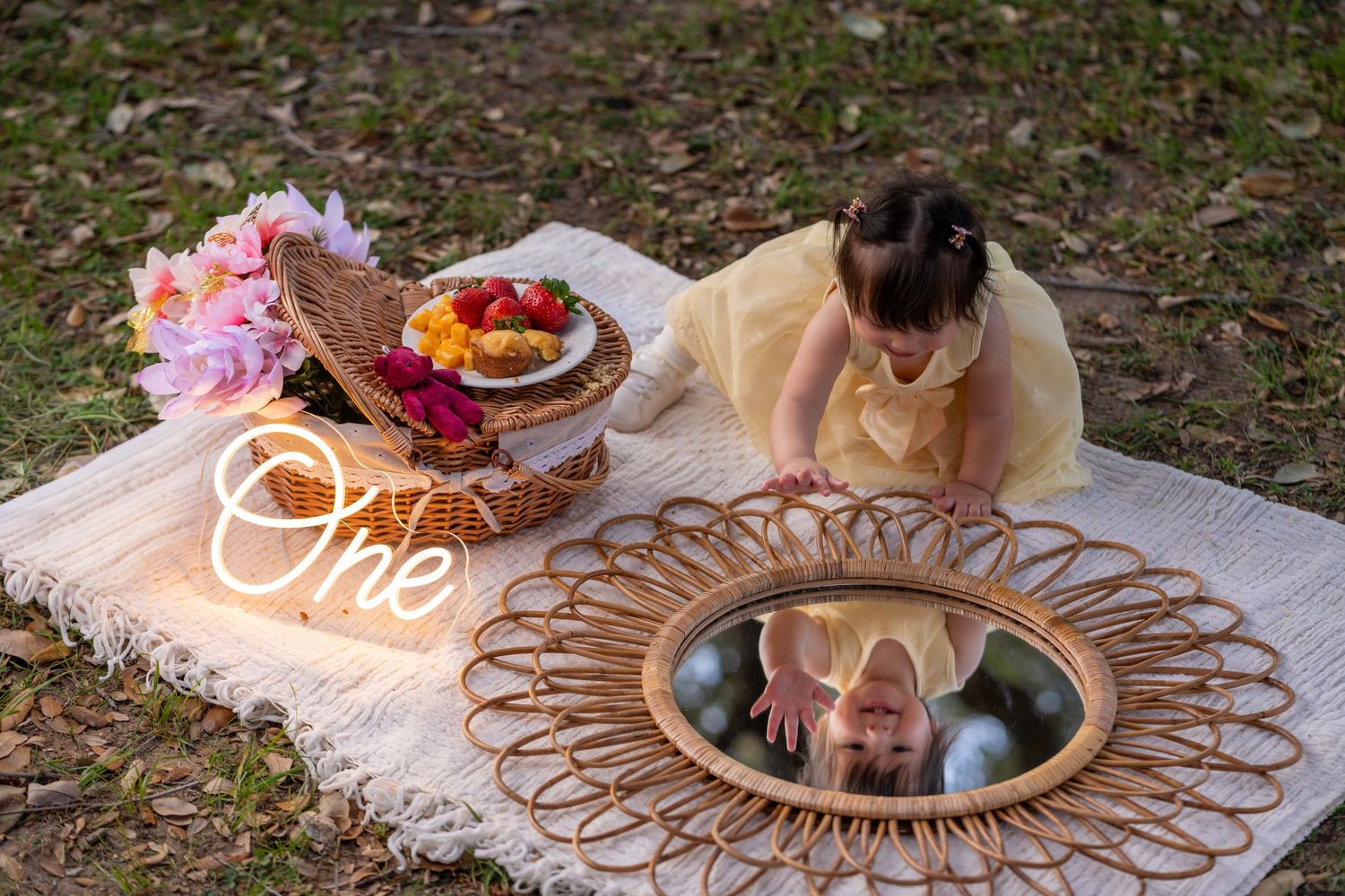 Toddler girl in yellow dress looks at mirror reflection during outdoor first birthday photoshoot with floral decor.