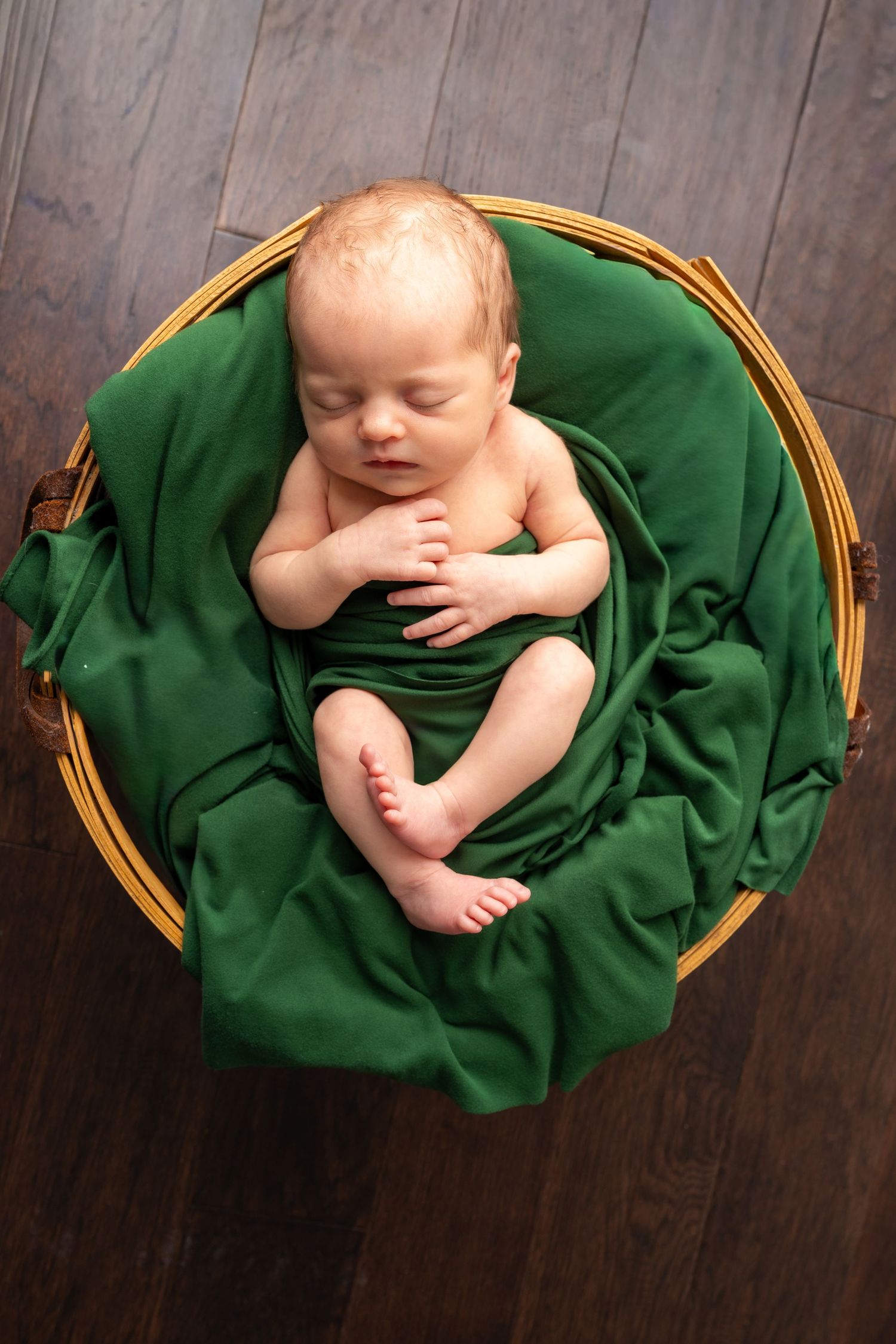Sleeping newborn baby nestled in a wicker basket with green fabric on a dark wood floor.