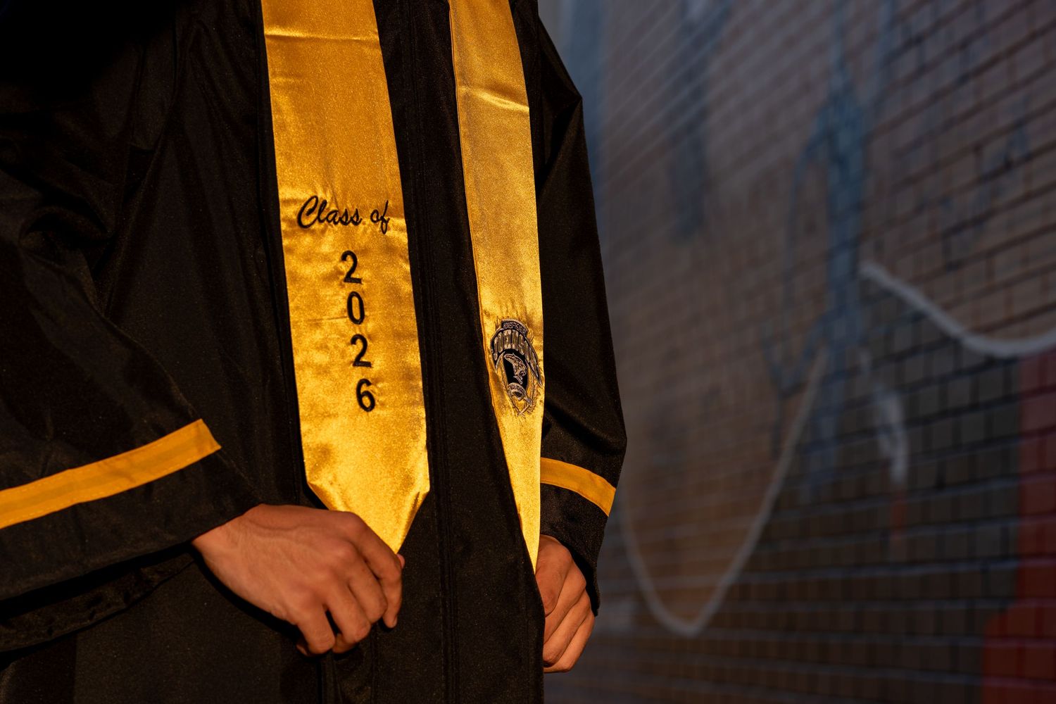 Person wearing black graduation gown with gold Class of 2026 stole, standing against a brick wall.