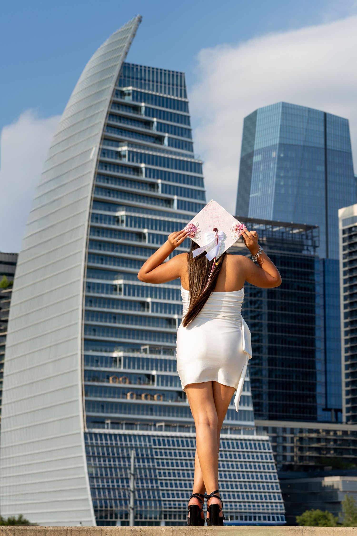 Woman in white dress holding graduation cap before modern city skyscrapers on a sunny day.
