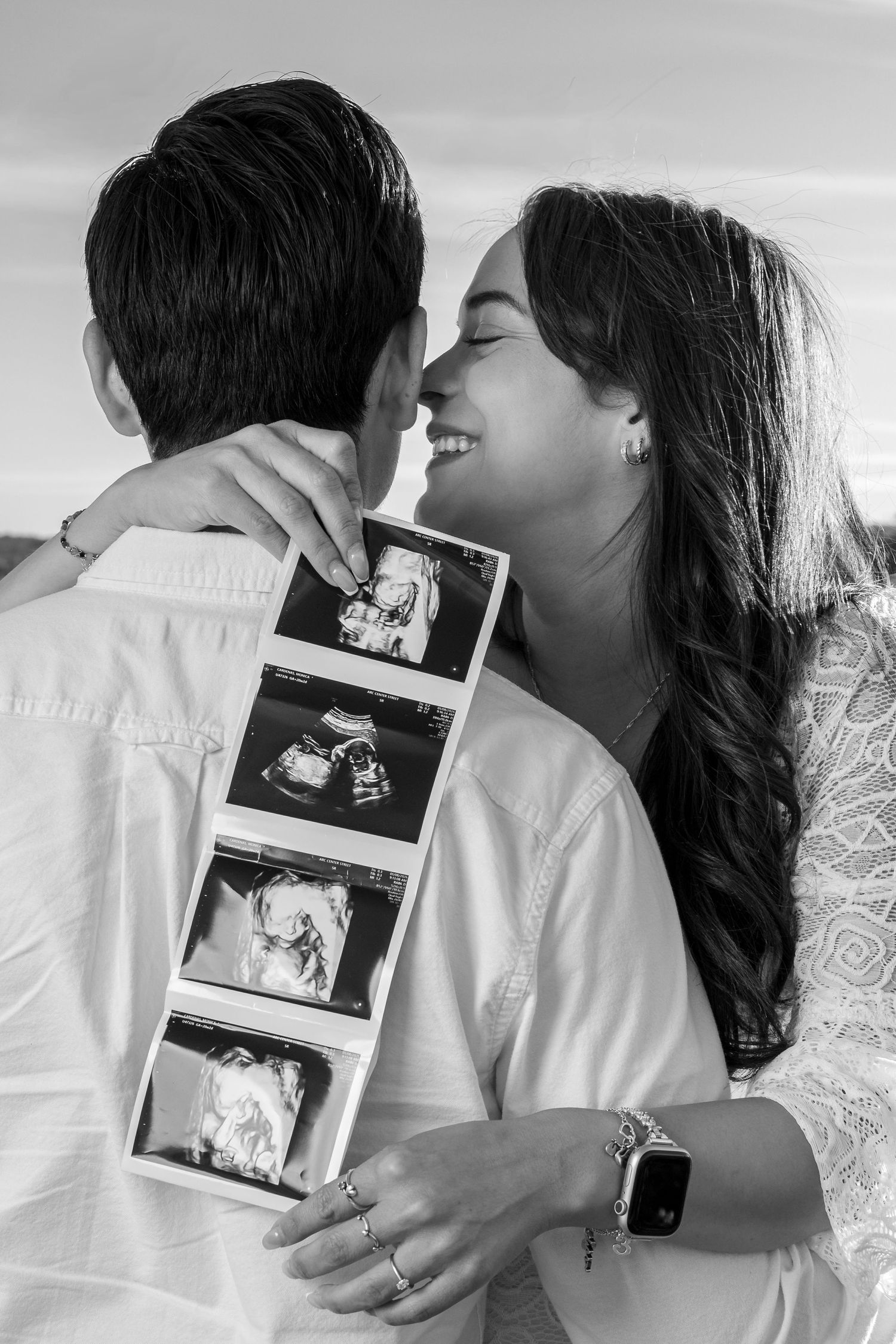 Black and white photo of a smiling couple embracing and holding ultrasound photos, announcing their pregnancy.