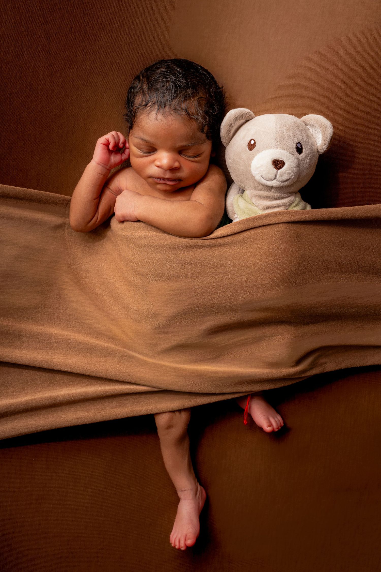 Sleeping newborn baby wrapped in brown fabric beside a teddy bear on a warm brown background.