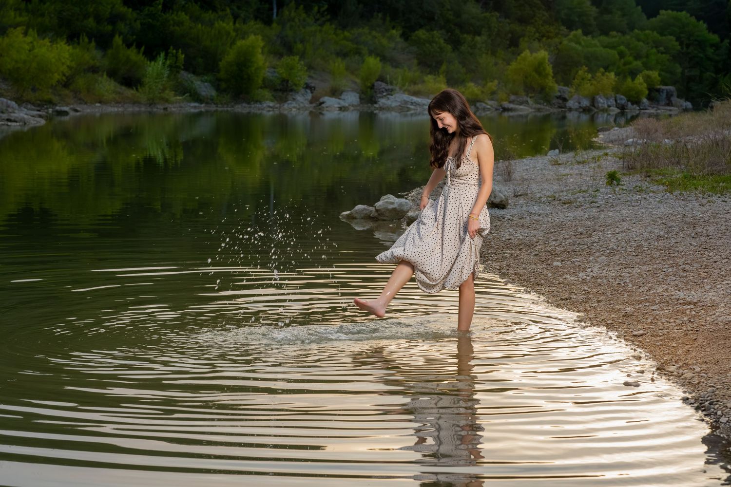 Young woman in a white dress wading in a calm lake, splashing water with her foot near a rocky shoreline.