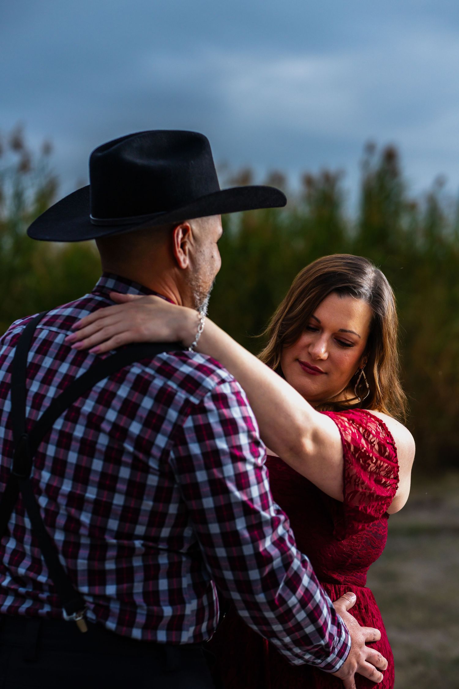A romantic couple embraces during a photoshoot, with the person in plaid and a cowboy hat holding their partner in red.