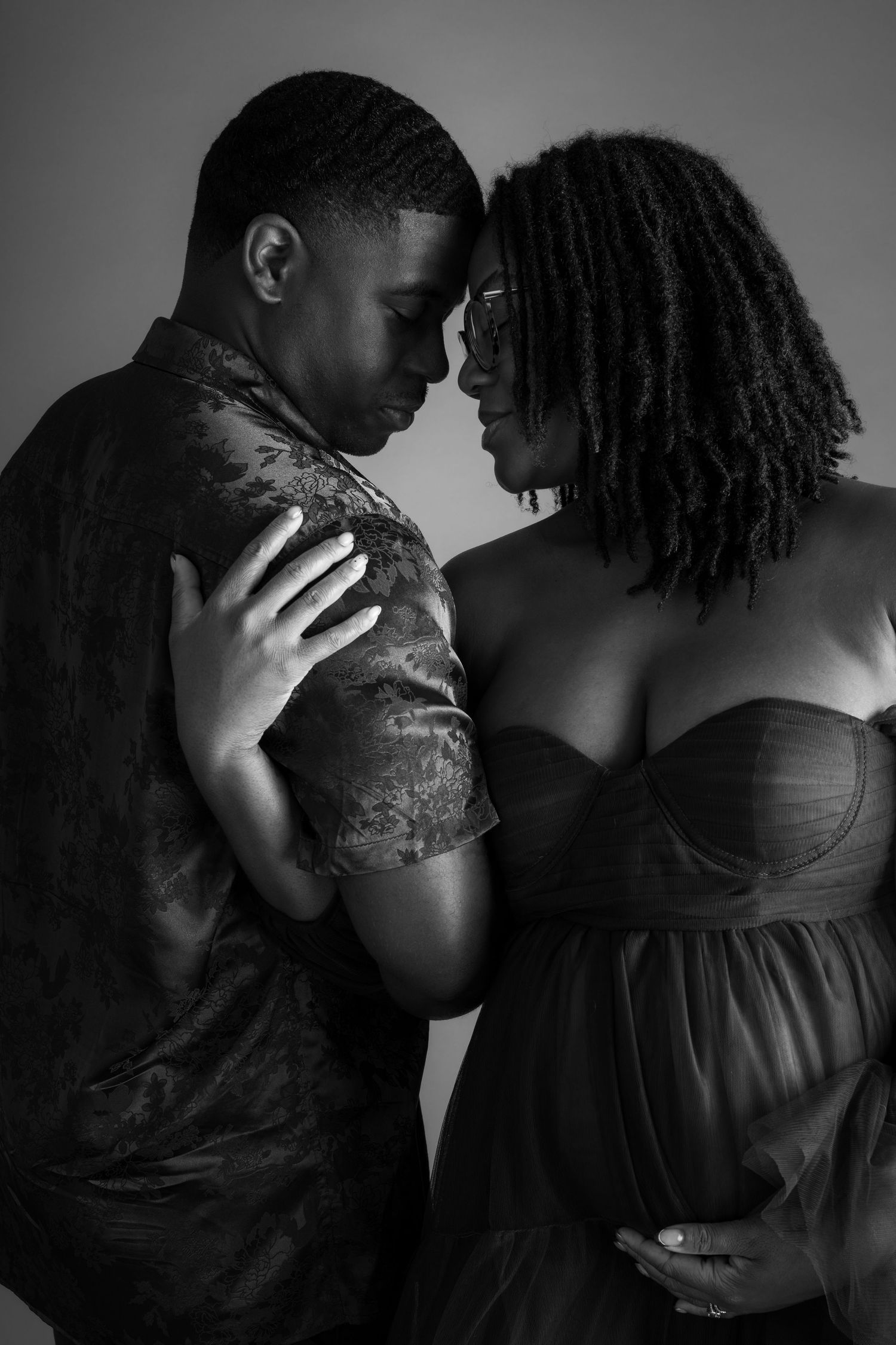 Black and white maternity photo of a couple tenderly touching foreheads, woman in strapless gown cradling baby bump.
