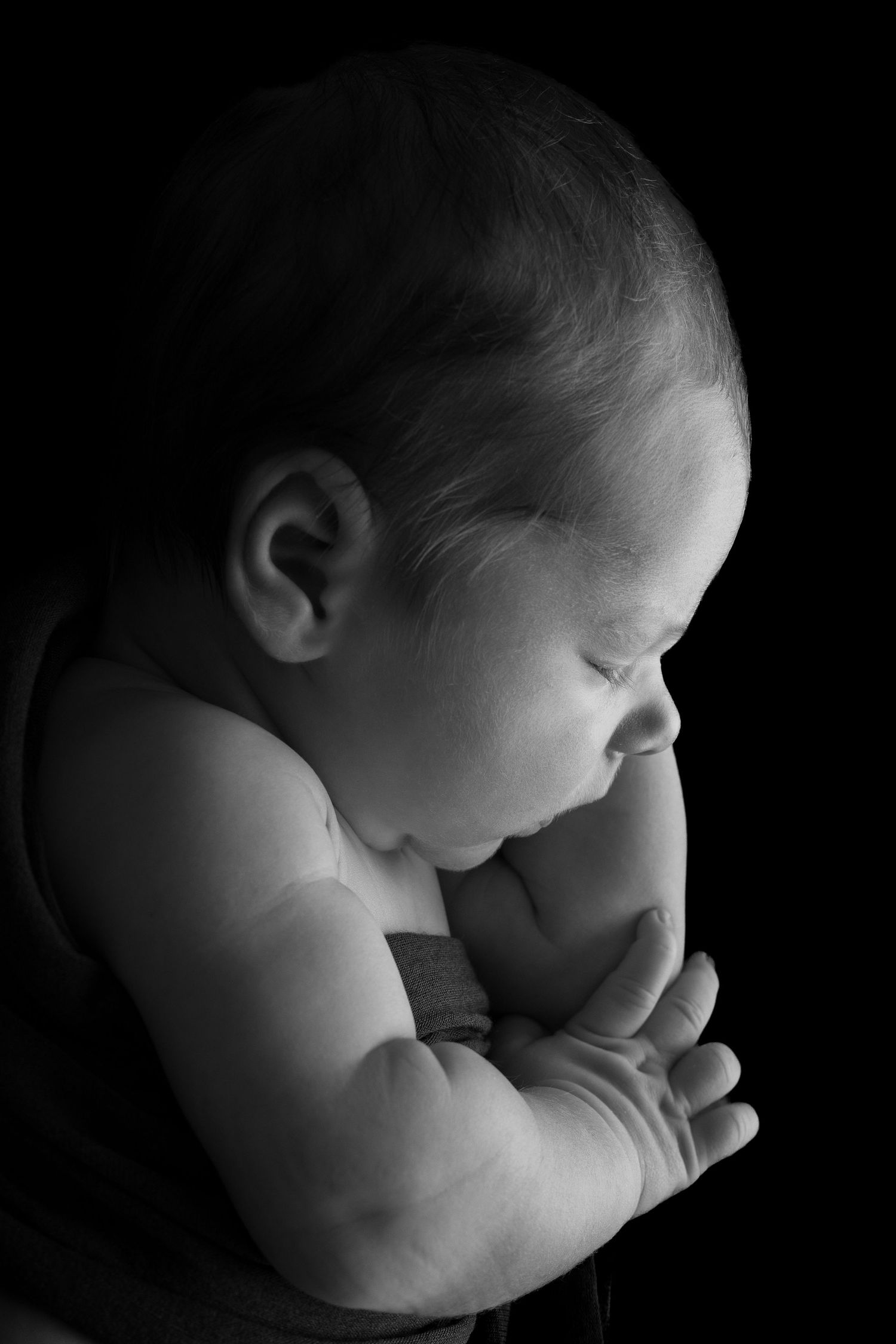Black and white newborn baby portrait with eyes closed, hands folded, against dark background.