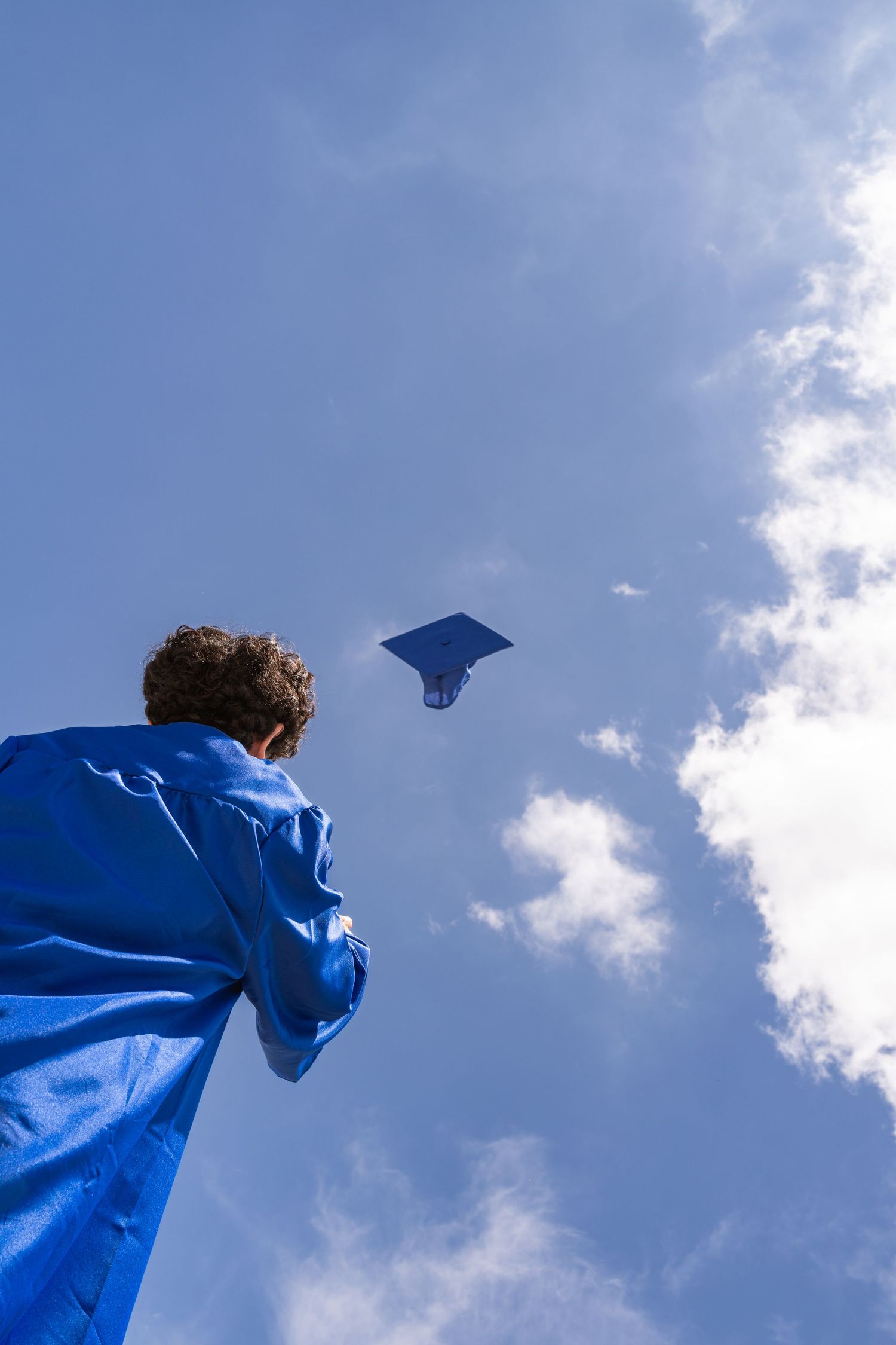 Graduate in blue gown tosses mortarboard cap into bright blue sky during outdoor graduation ceremony.