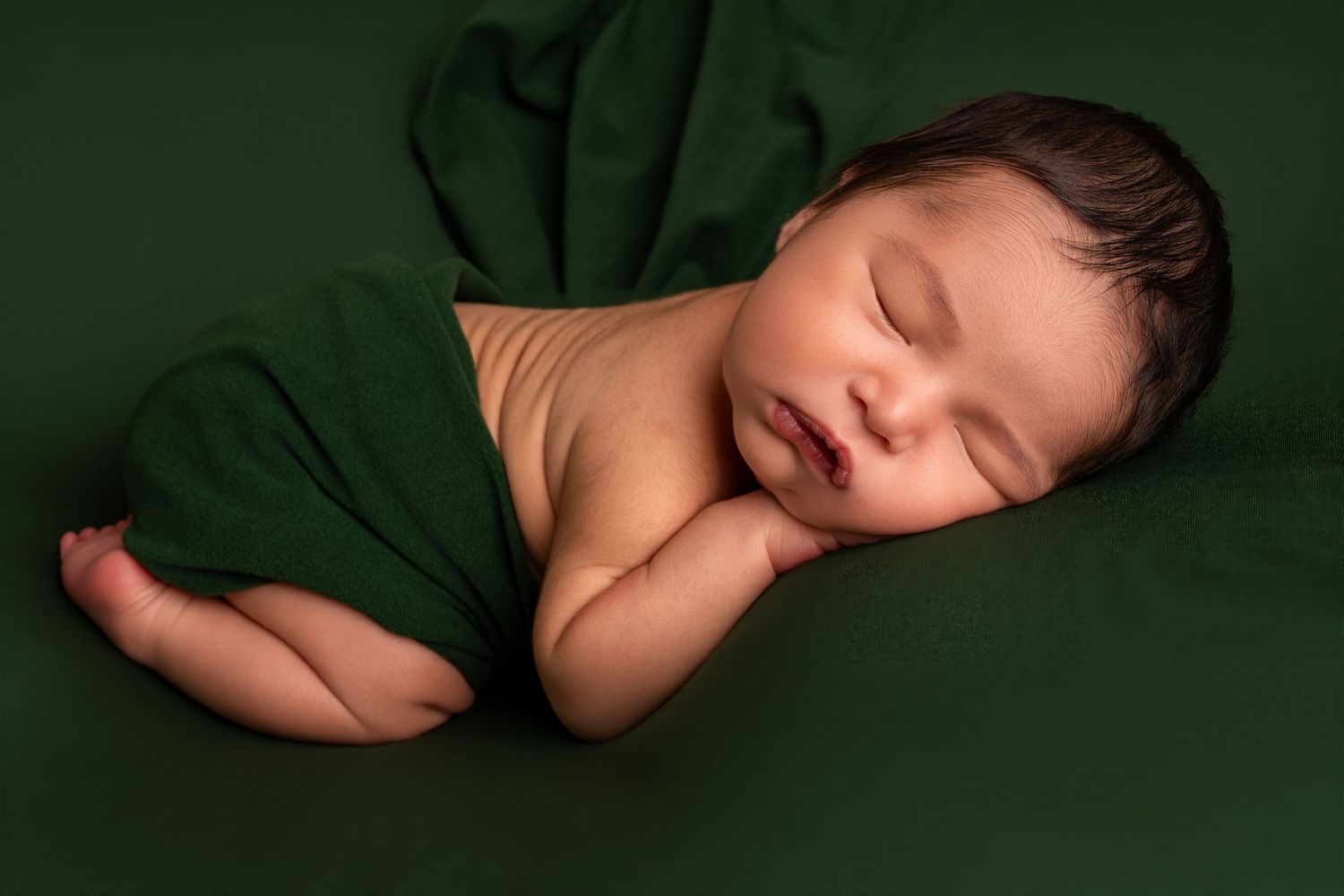 Sleeping newborn baby with dark hair rests on a dark green blanket, wrapped in matching green fabric, in a peaceful newborn portrait.