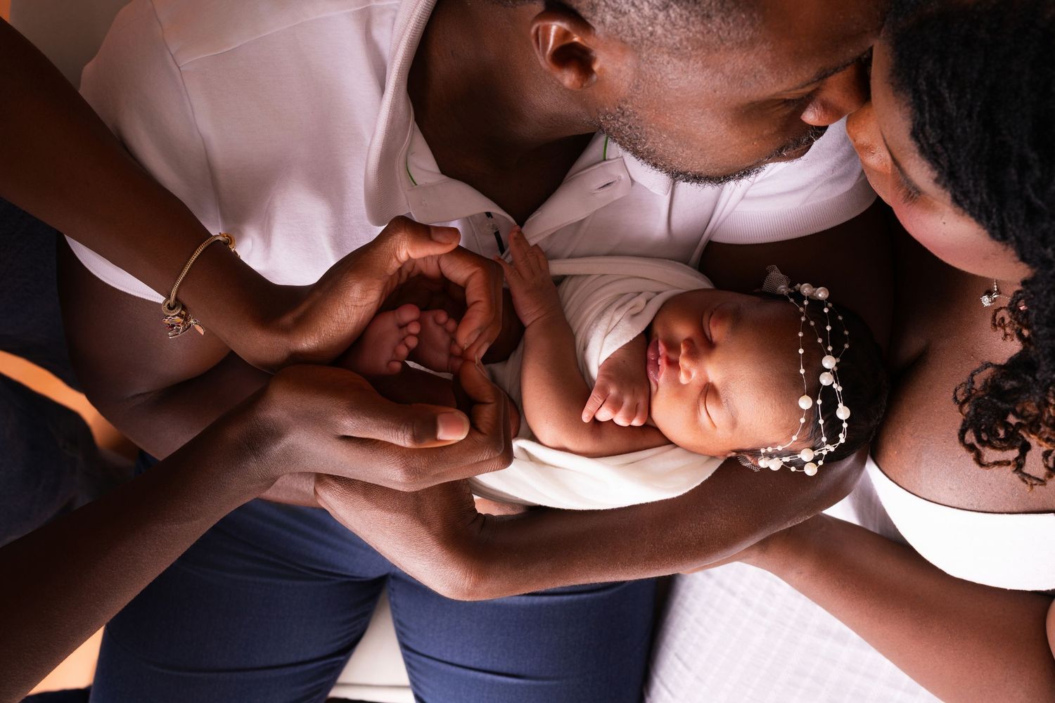 Two parents gently cradle a sleeping newborn baby wearing a beaded headband in a tender overhead family portrait.