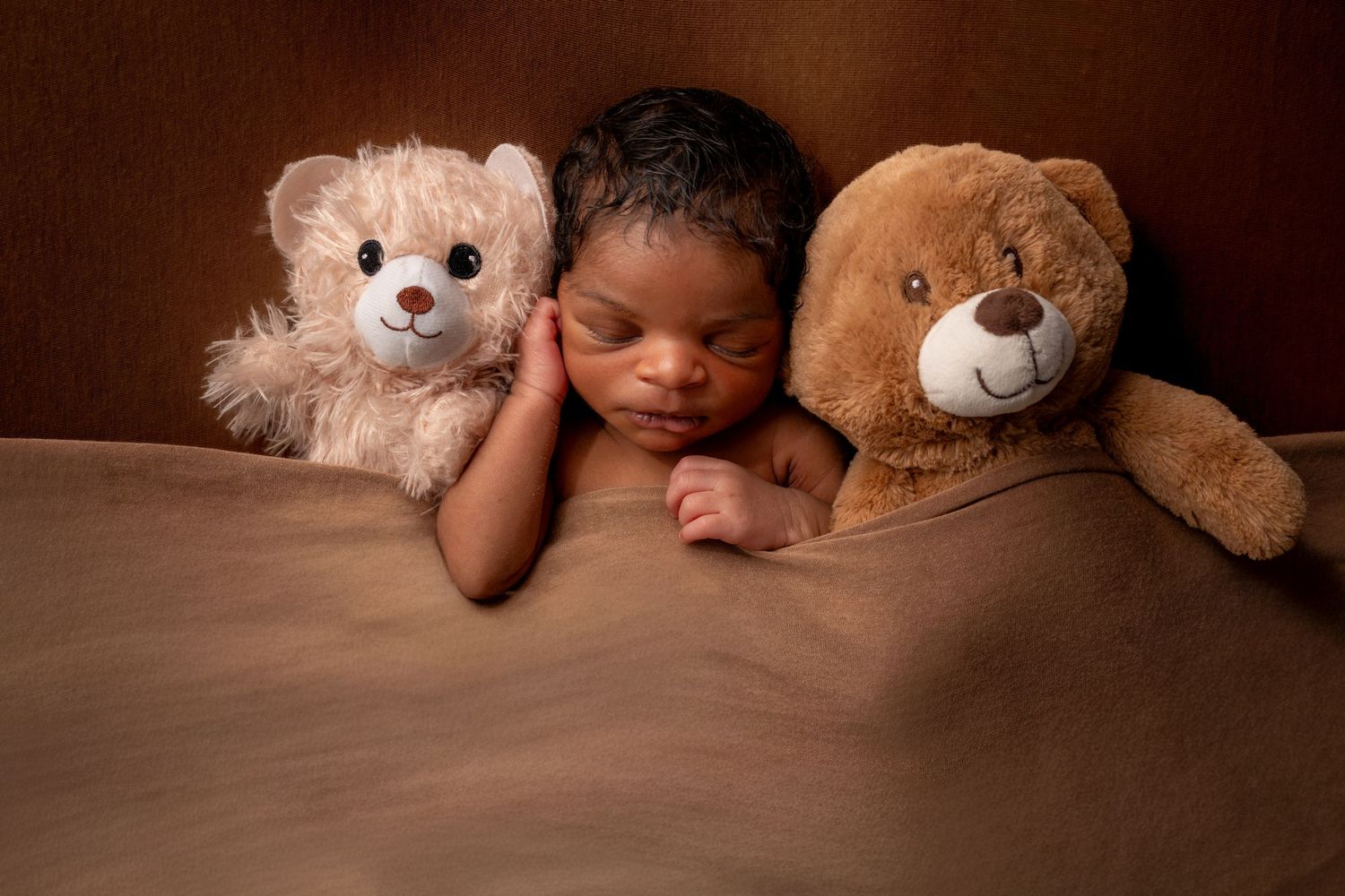 Sleeping newborn baby nestled between two teddy bears on a brown blanket in a cozy studio portrait.