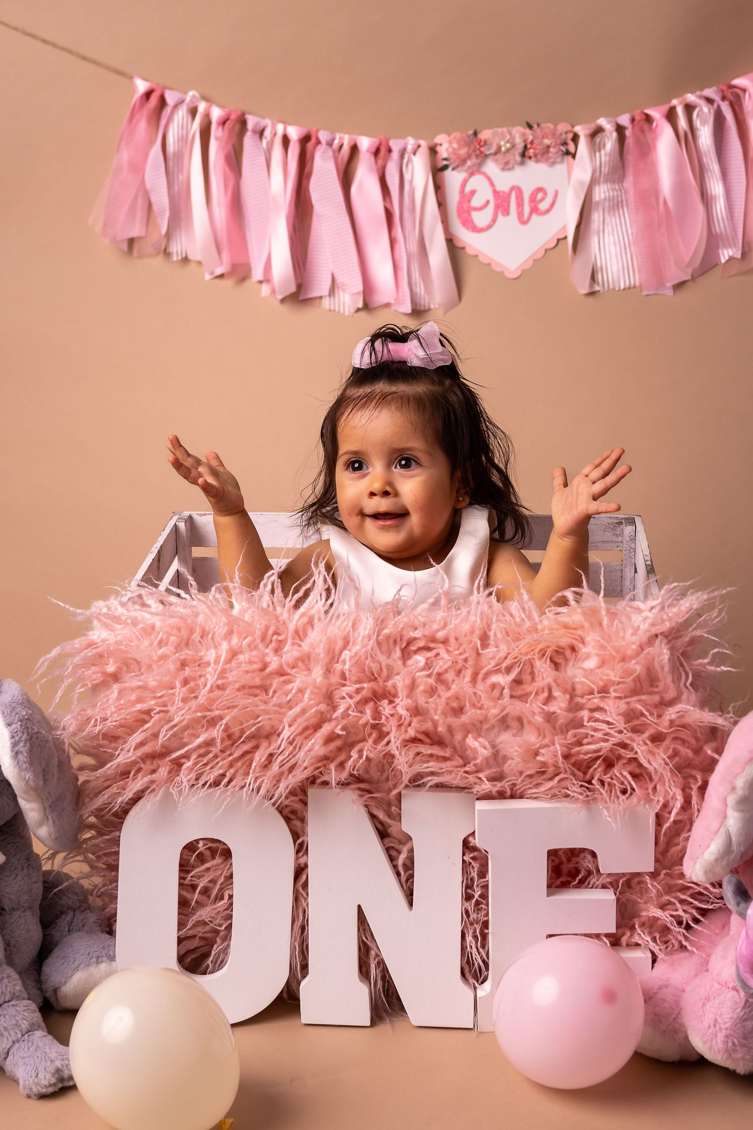 Happy baby girl in pink fluffy dress celebrating first birthday with ONE letters, balloons, and pink banner.