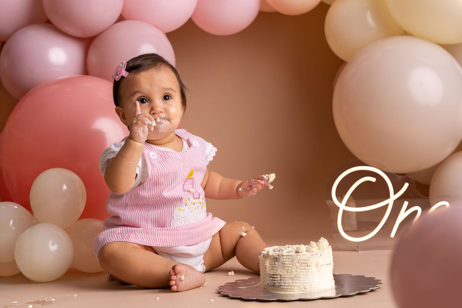Baby girl in pink outfit smashes first birthday cake surrounded by pink and white balloons on beige backdrop.