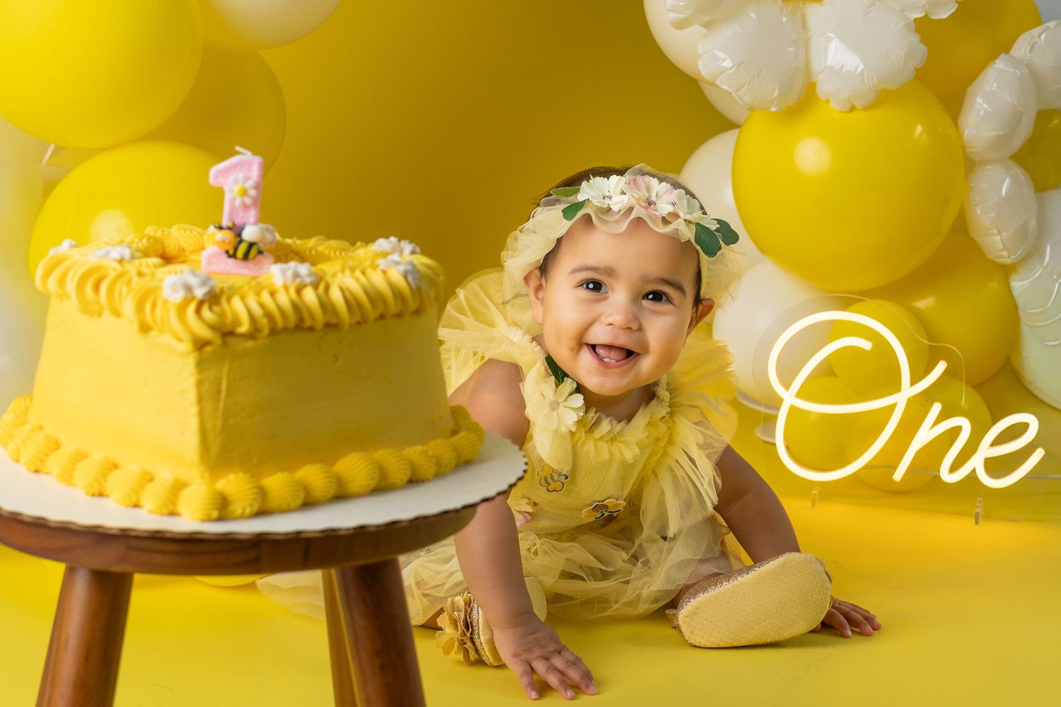 Baby girl in yellow dress smiles beside a yellow first birthday cake, surrounded by yellow balloons.