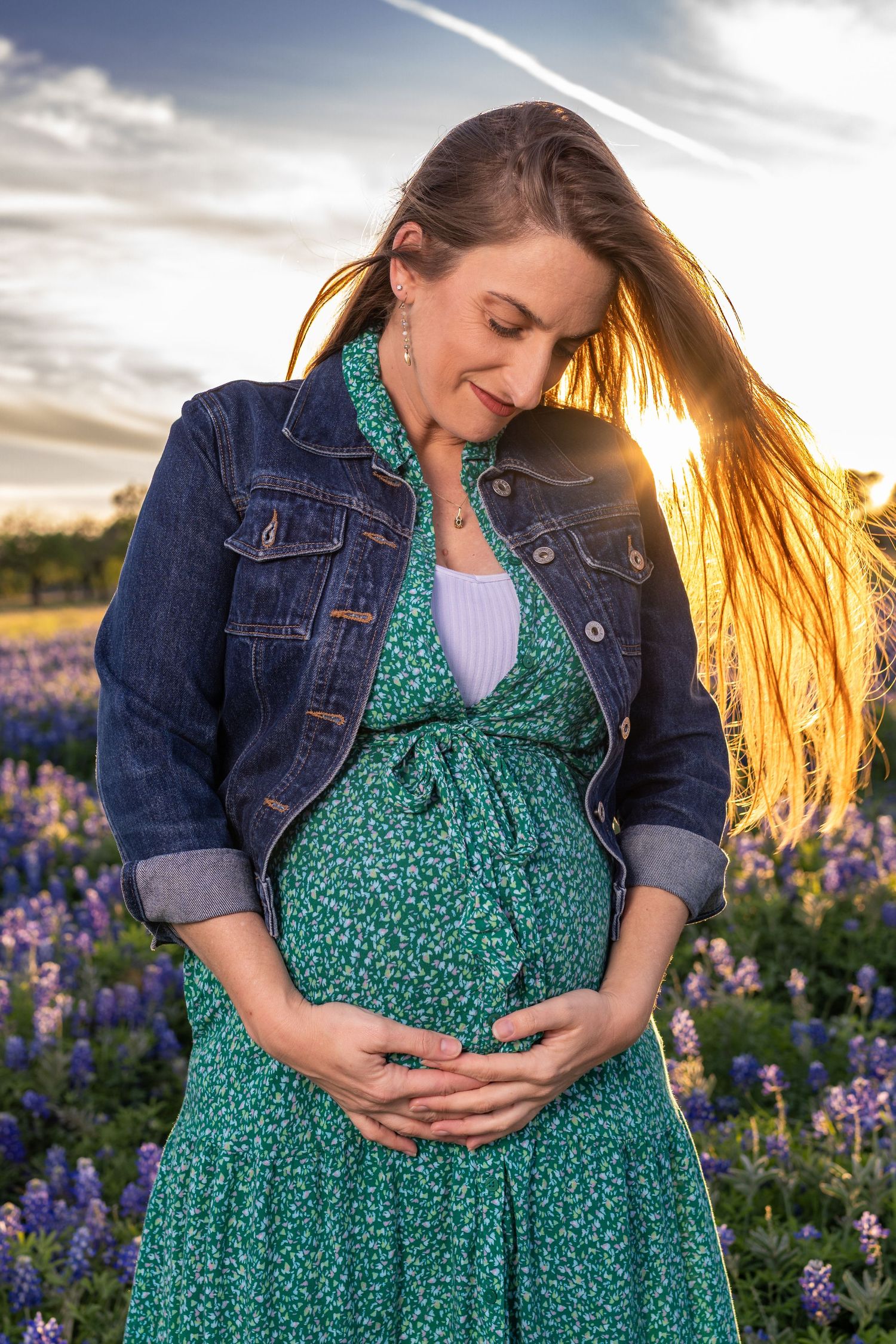 Pregnant woman in green floral dress and denim jacket cradling baby bump in a bluebonnet field at golden sunset.