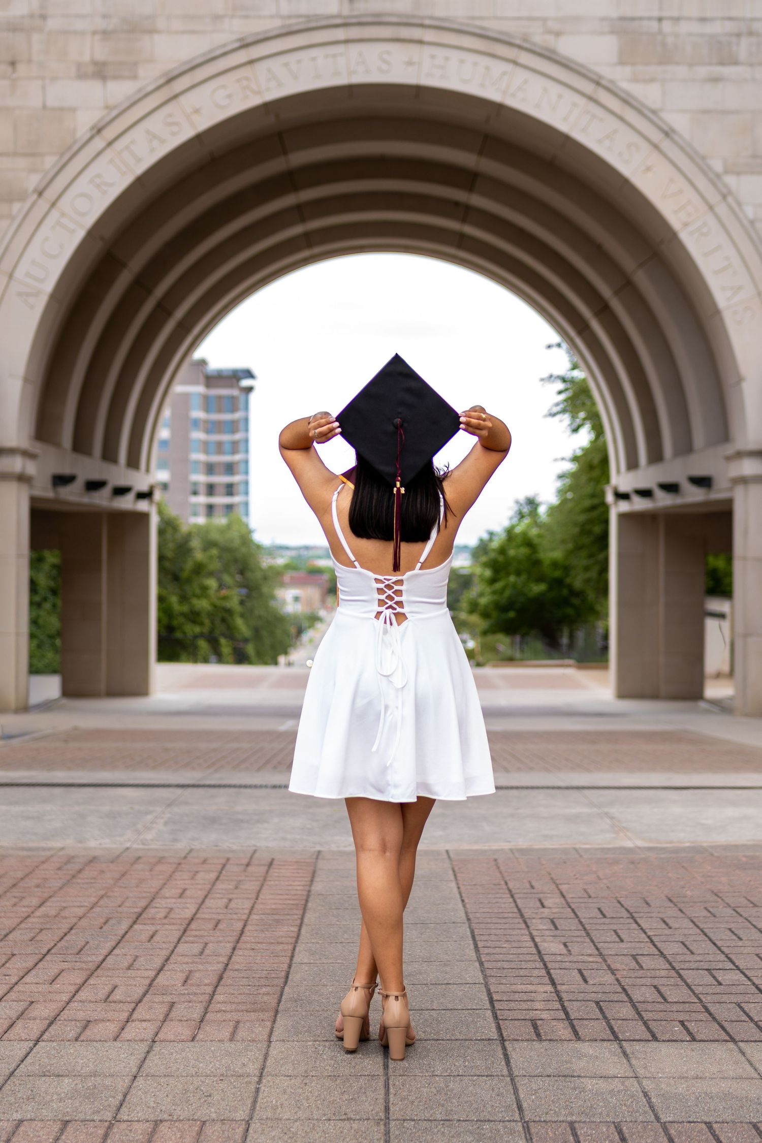 Graduate in white dress holds mortarboard cap under stone arch, viewed from behind, celebrating college graduation outdoors.