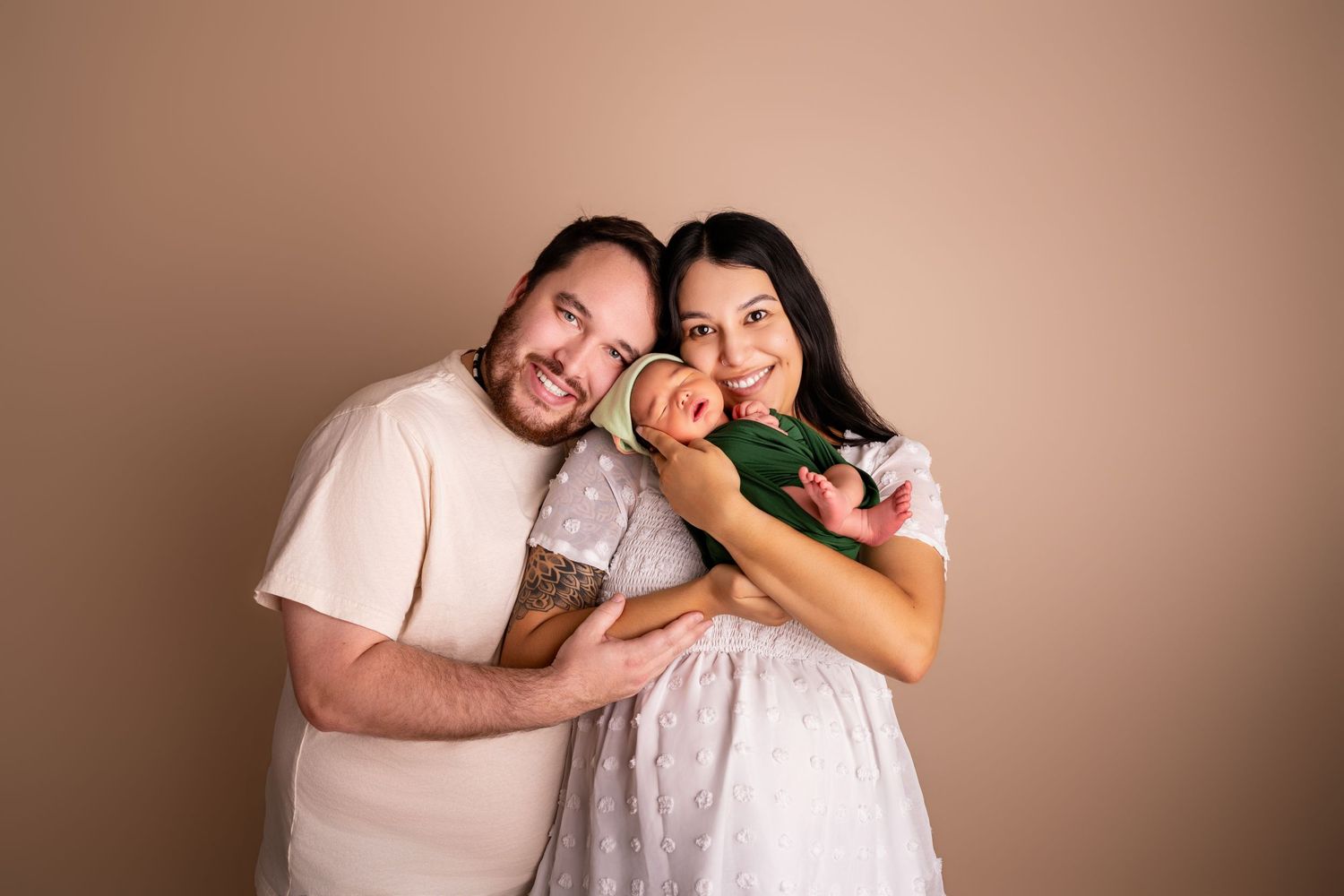 Smiling couple in a cream shirt and white dress cuddle a newborn baby in a green outfit in a warm studio family portrait.
