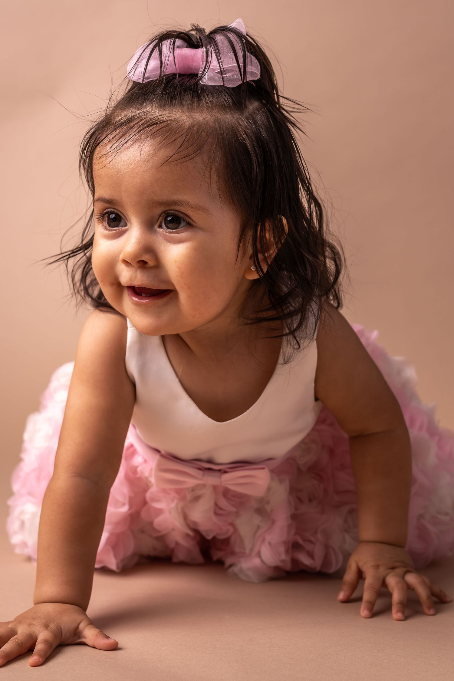 Smiling baby girl in white top and pink tutu skirt crawling on beige background.