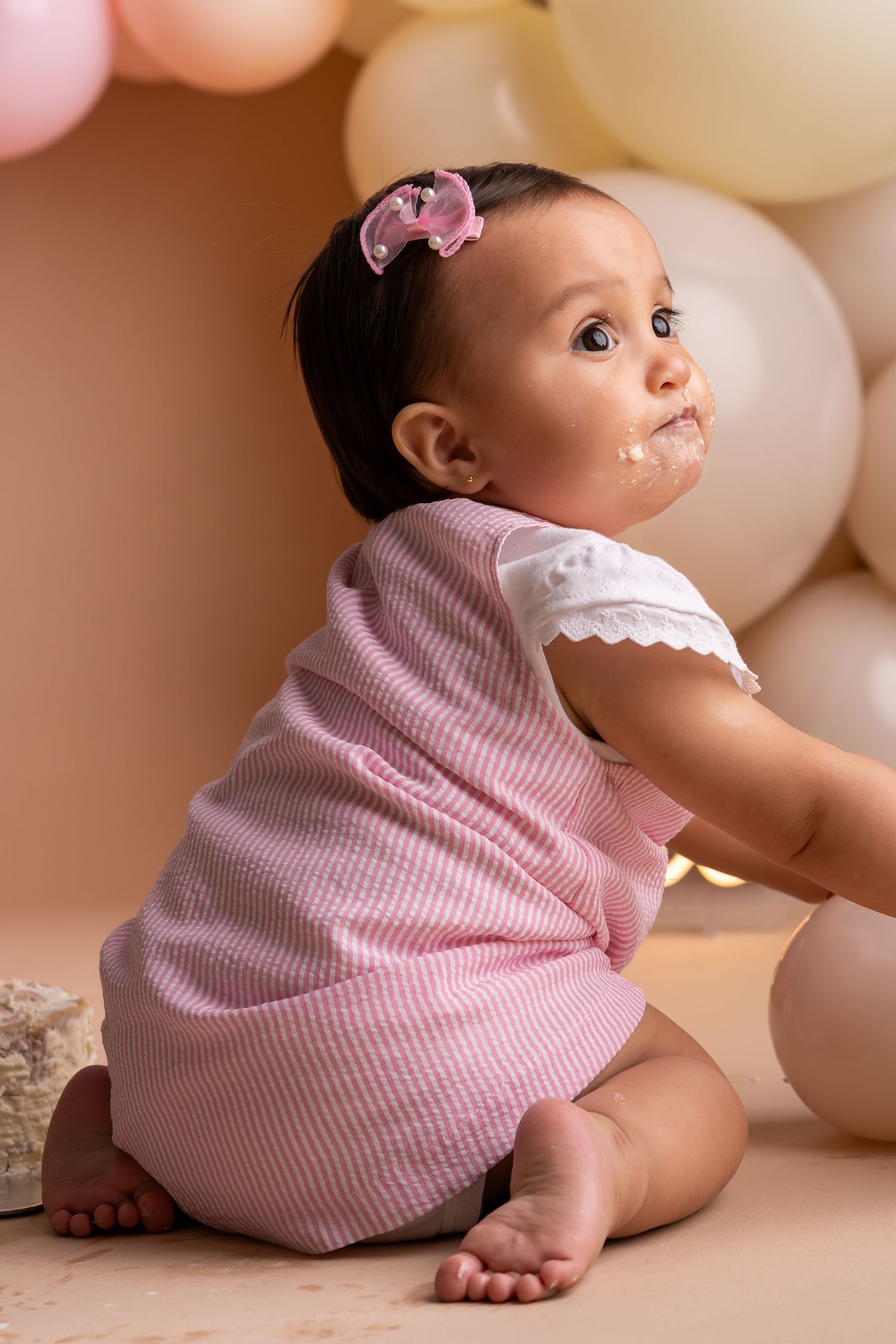 Baby in pink gingham dress poses against beige backdrop with balloons for first birthday photoshoot.